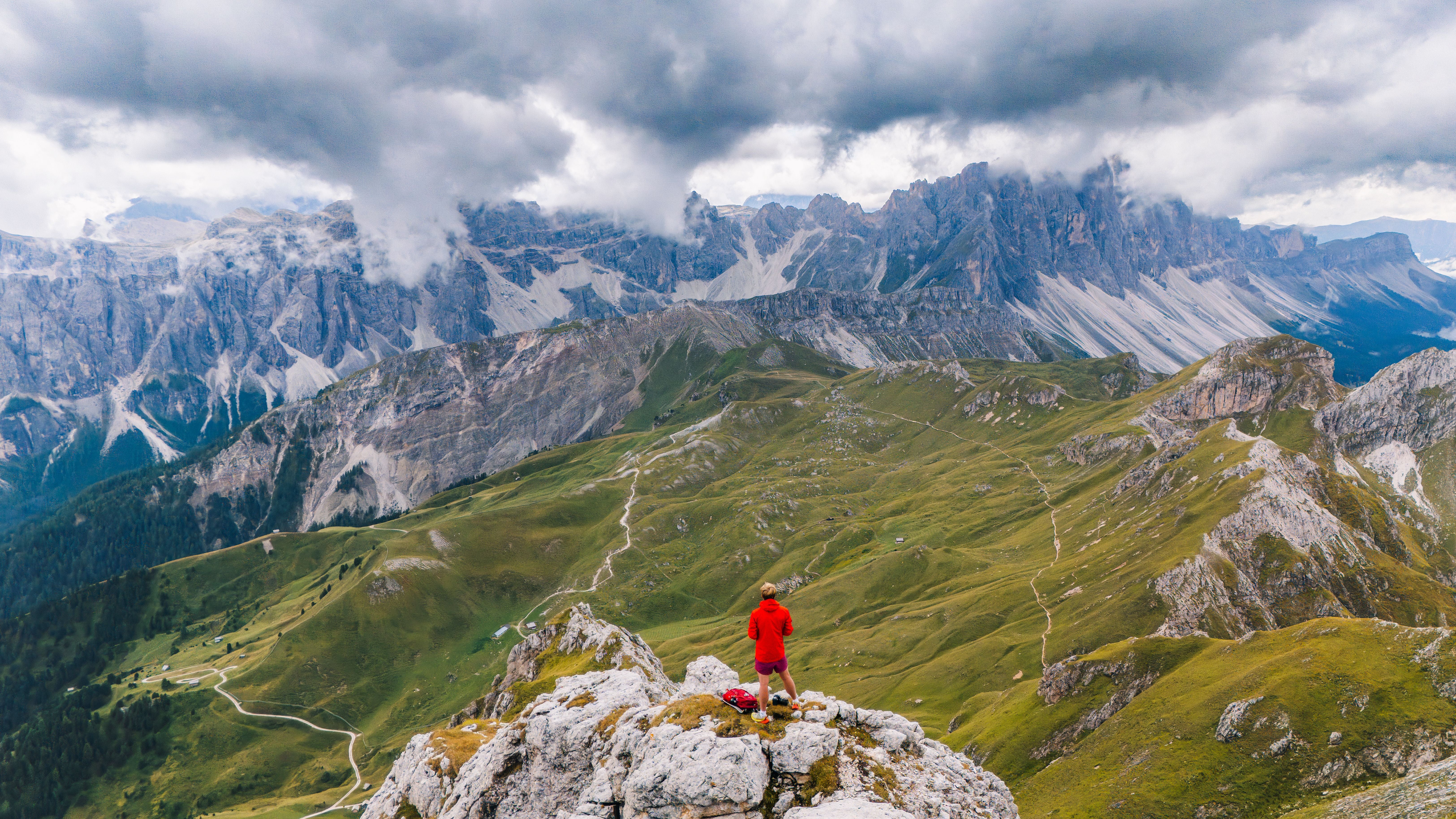 Person in roter Jacke steht auf Felsen mit Blick über grüne Dolomitenlandschaft und dramatische Wolken.