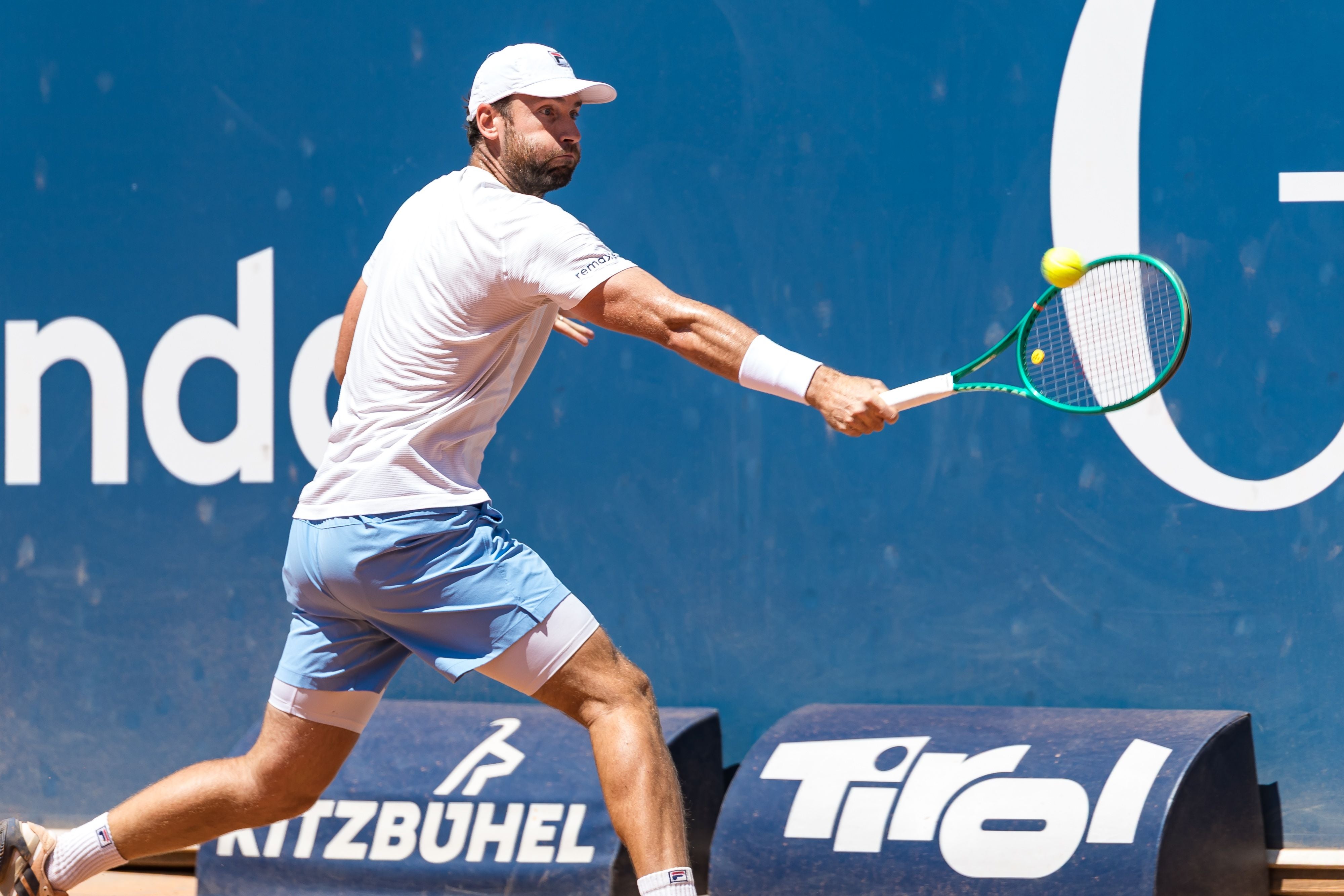 Ein Tennisspieler schlägt auf Sandplatz einen Ball mit dem Schläger, im Hintergrund Sponsorenlogos.