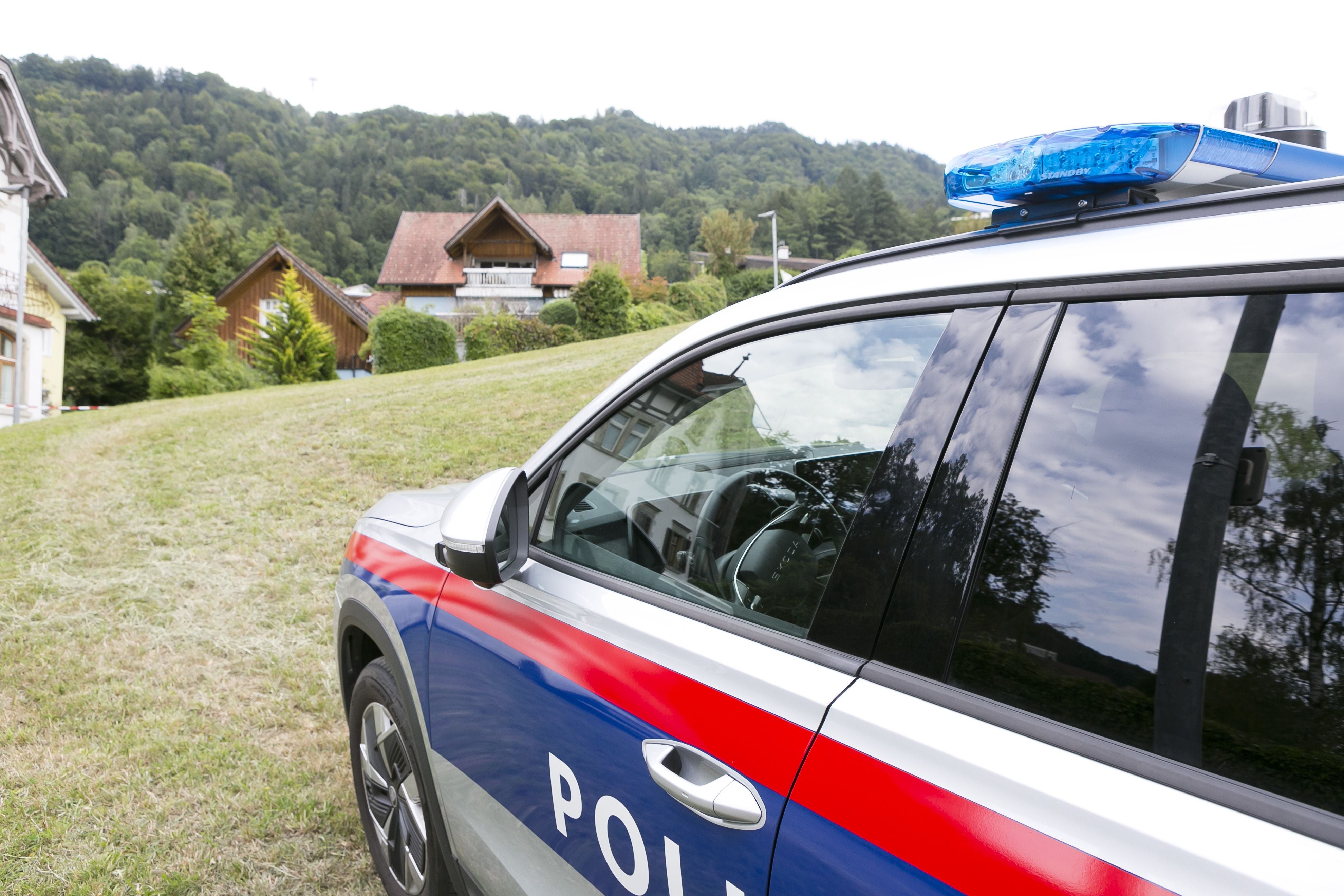Ein österreichisches Polizeiauto steht auf einer Wiese vor Wohnhäusern am Waldrand.