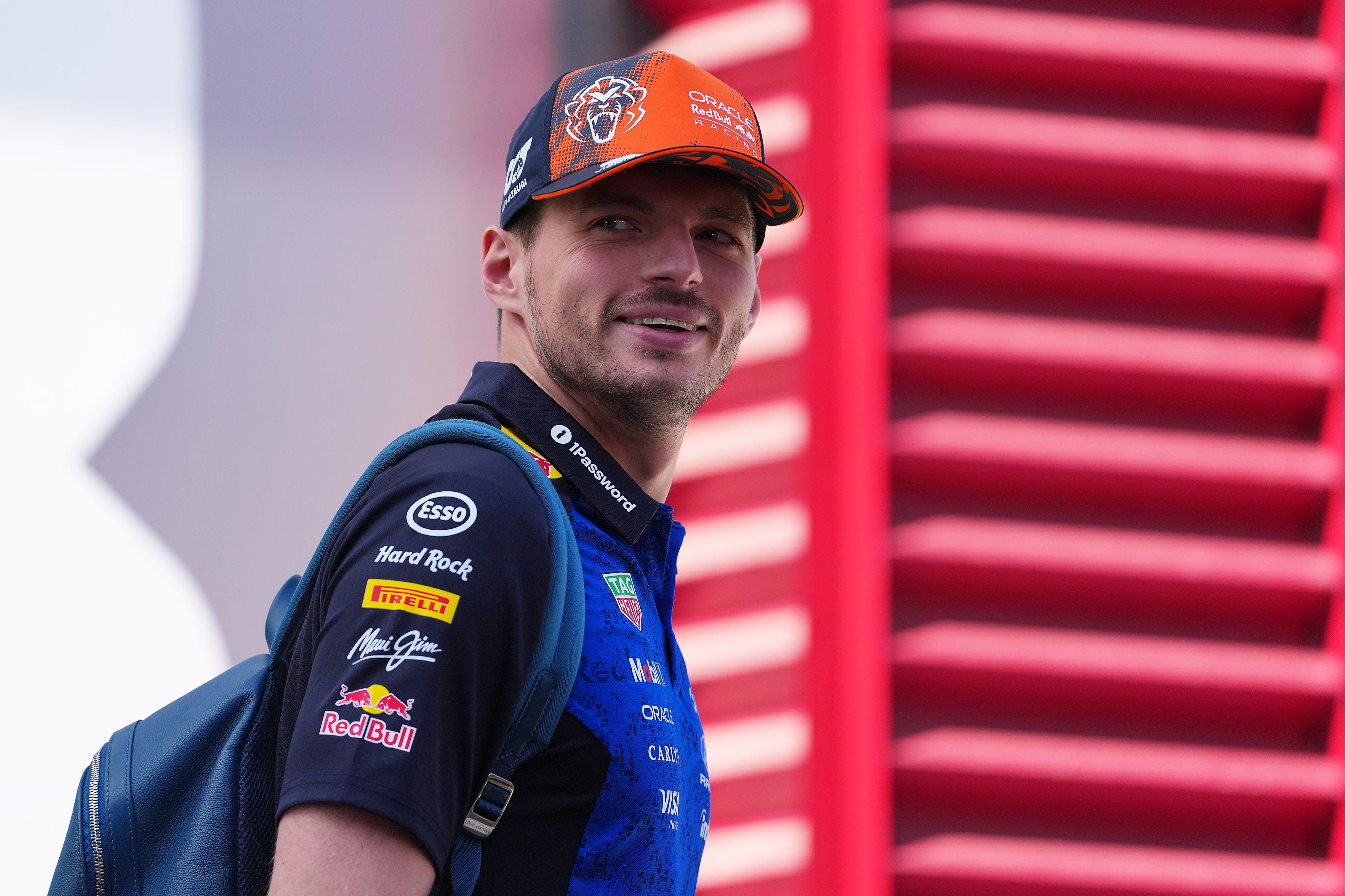 Ein Mann in Red-Bull-Teamkleidung und Cap steht lächelnd mit einem Rucksack vor rotem Hintergrund.