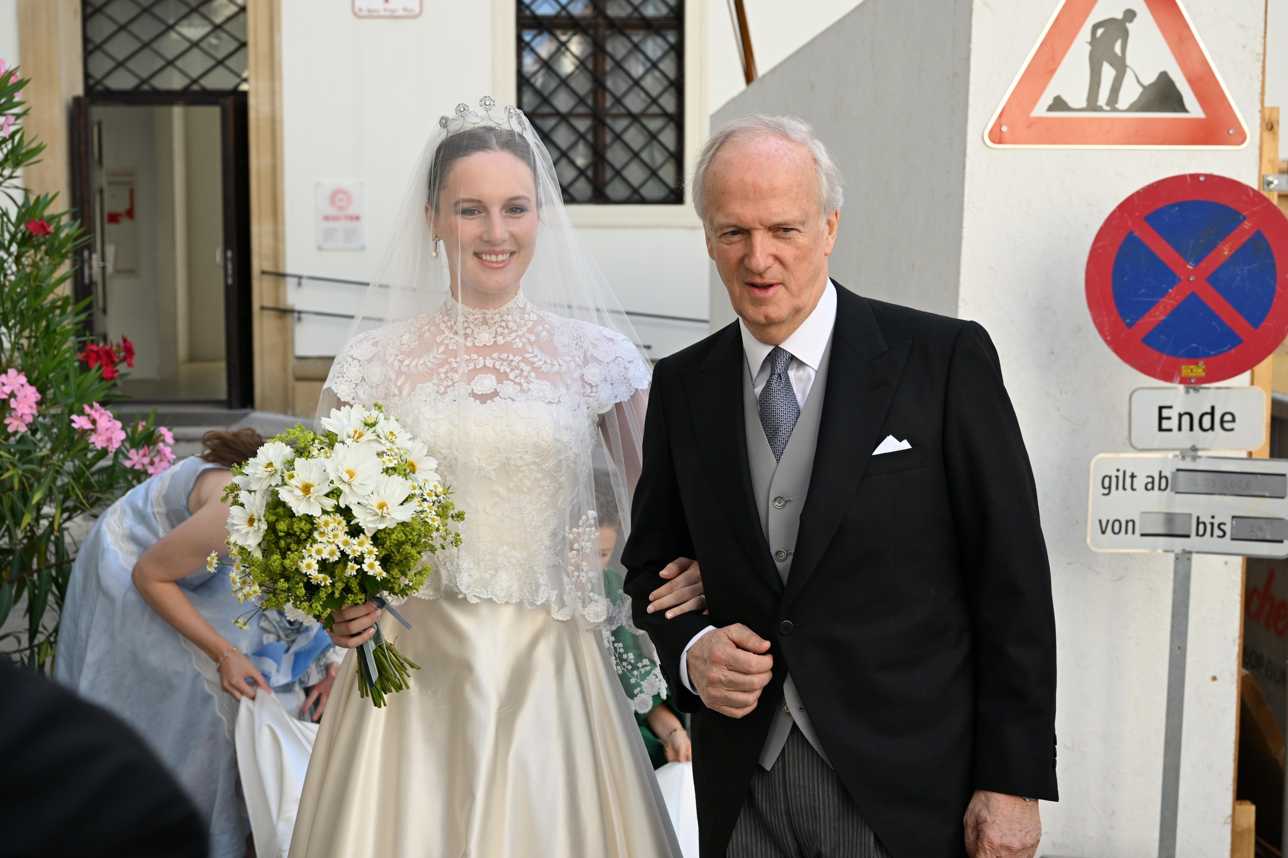 Braut im weißen Kleid mit Blumenstrauß und älterer Mann im Anzug bei einer Hochzeit draußen.