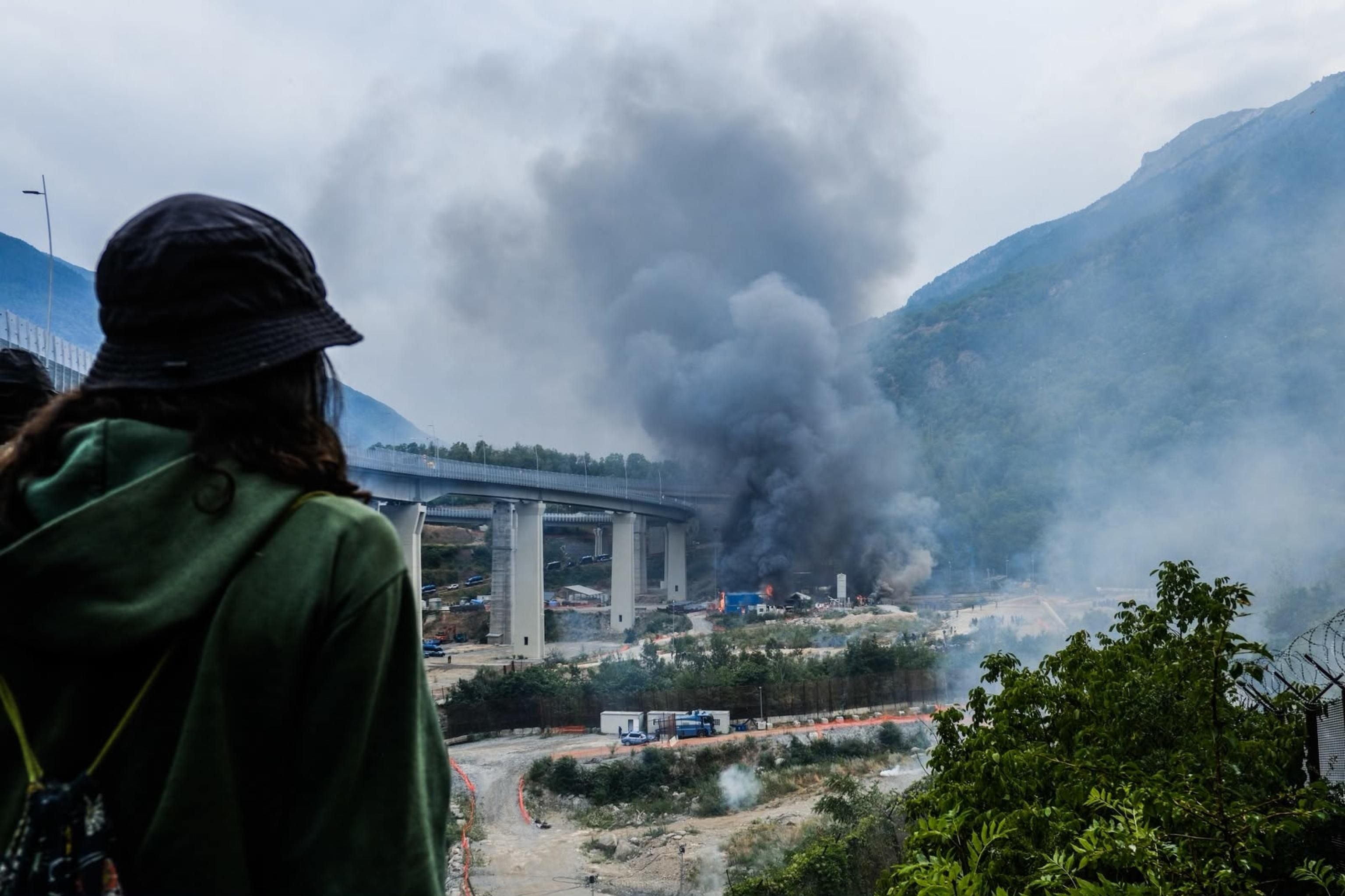 Eine Person blickt auf eine Baustelle, von der dichter Rauch aufsteigt; im Hintergrund Berge.