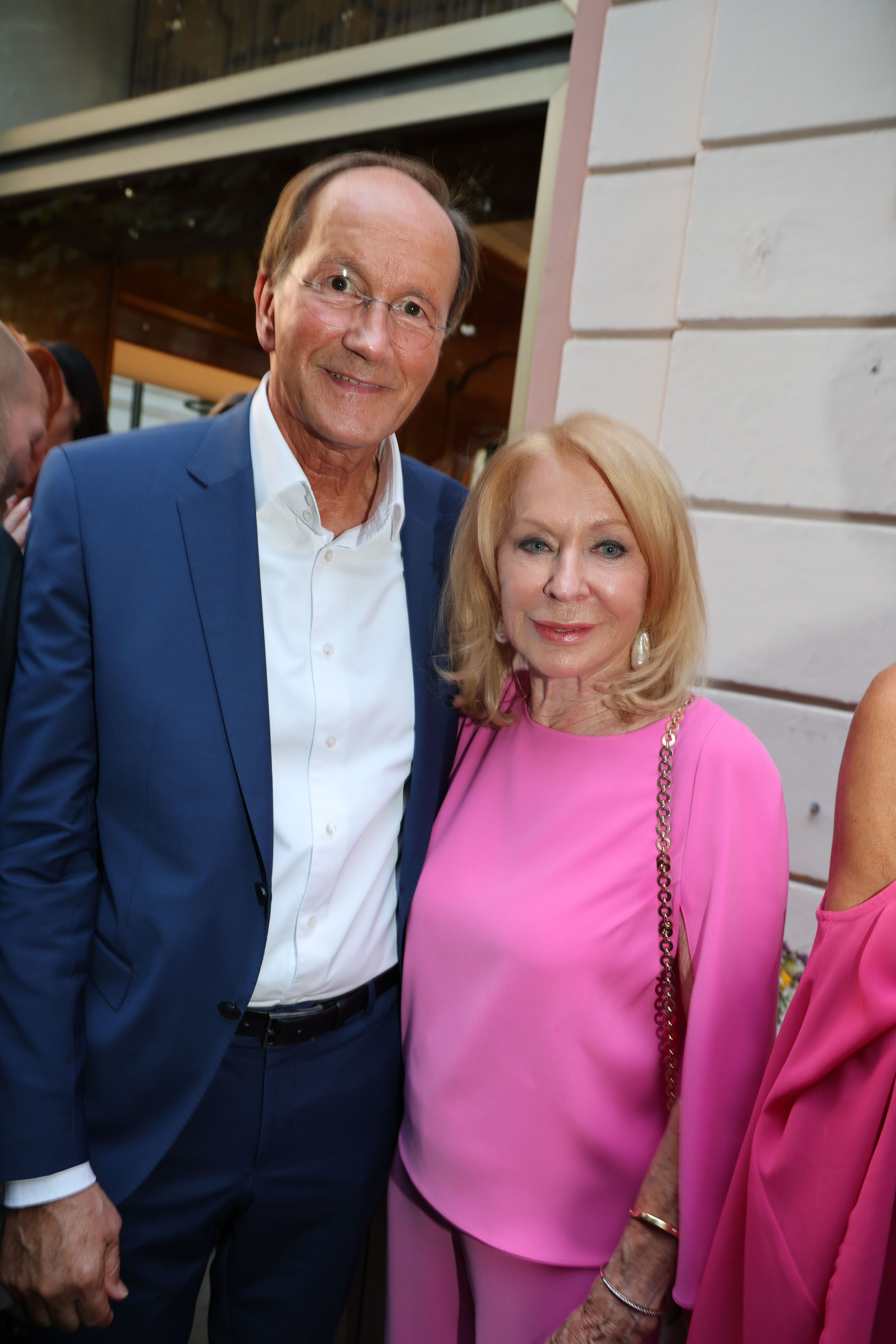 Ein älteres Paar lächelt, der Mann trägt einen blauen Anzug, die Frau ein pinkes Outfit.