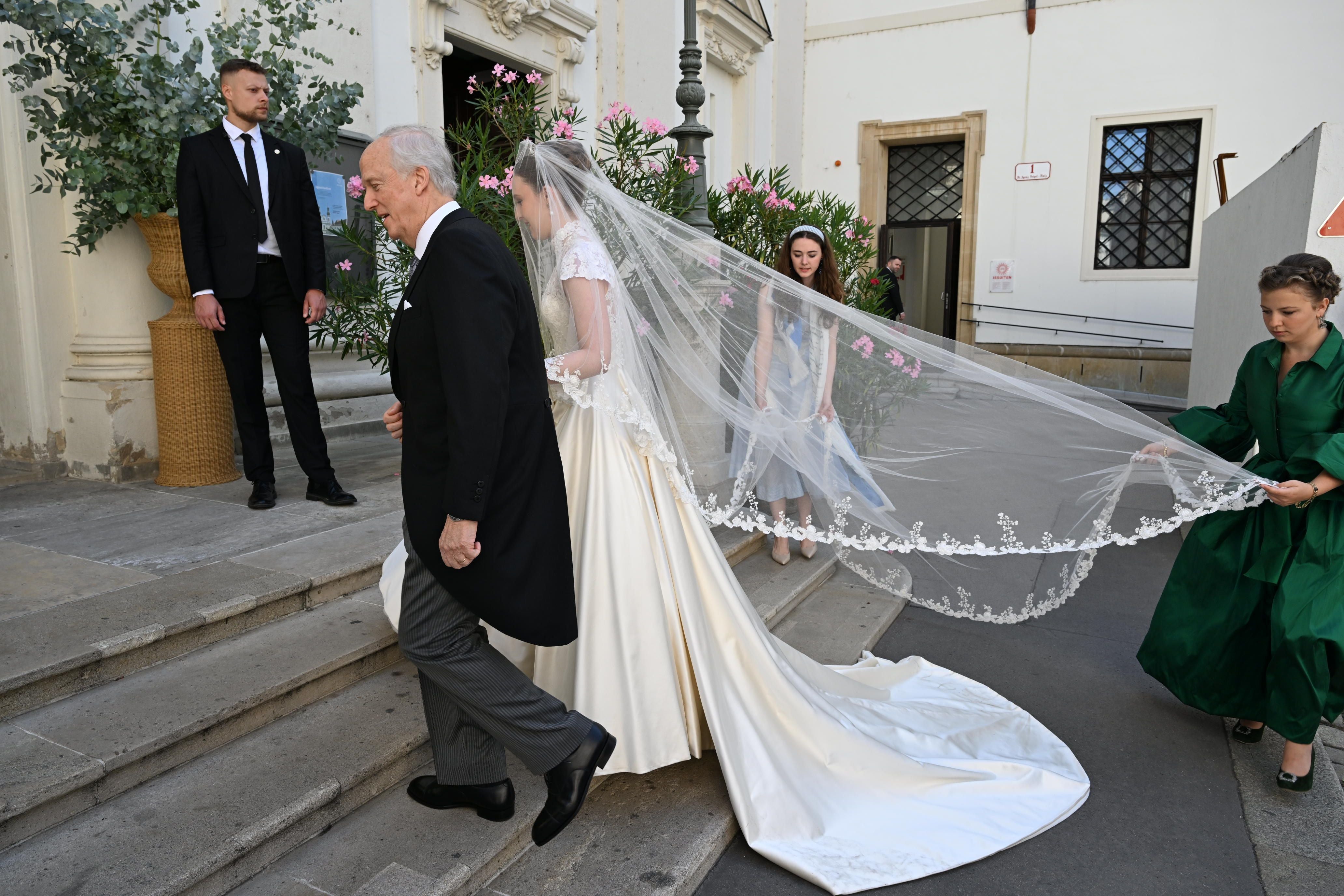 Eine Braut mit langem Schleier wird zur Hochzeit von Begleitern und einem älteren Herrn begleitet.