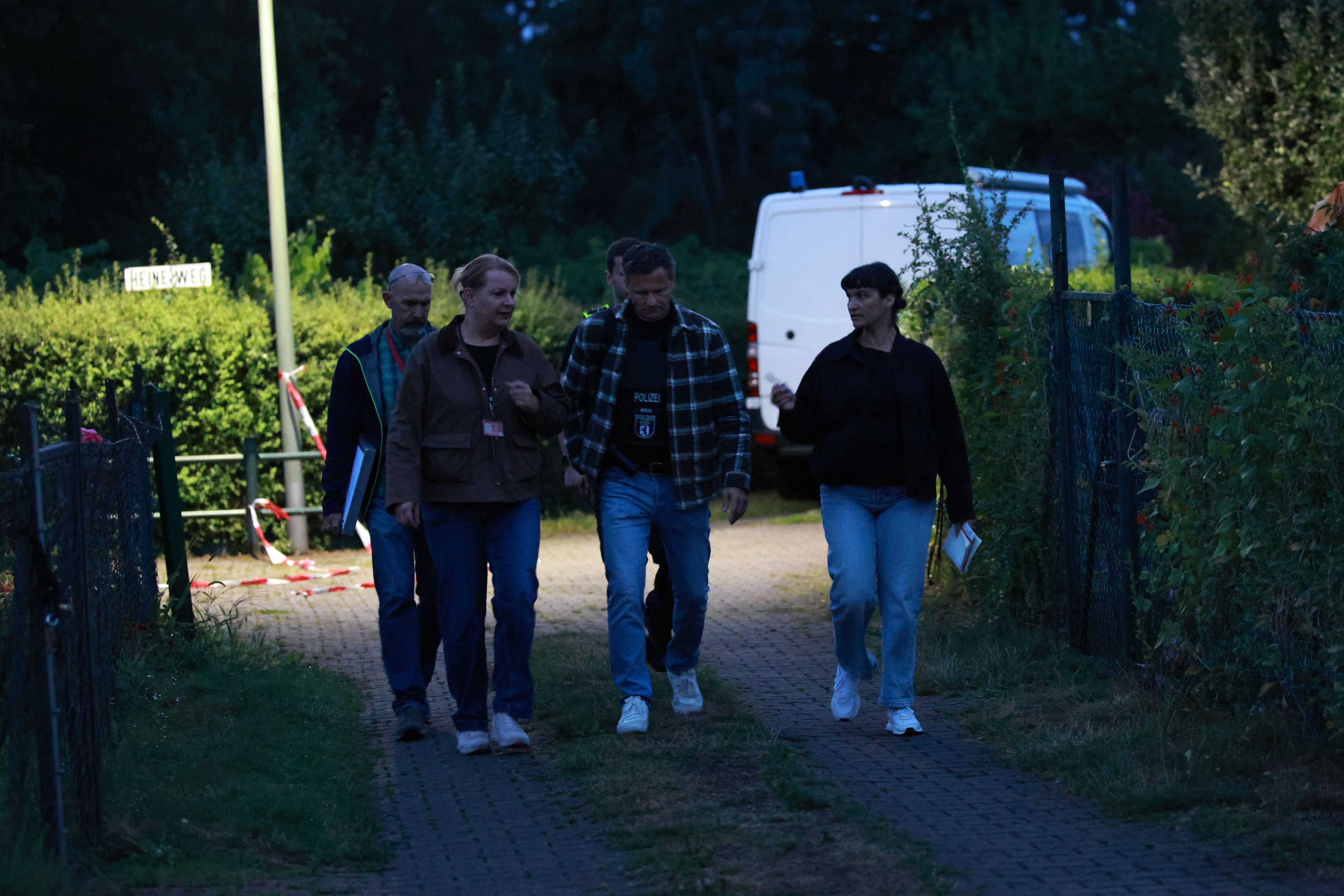 Ermittler gehen abends durch eine Gartenanlage in Berlin, im Hintergrund steht ein Polizeiwagen.