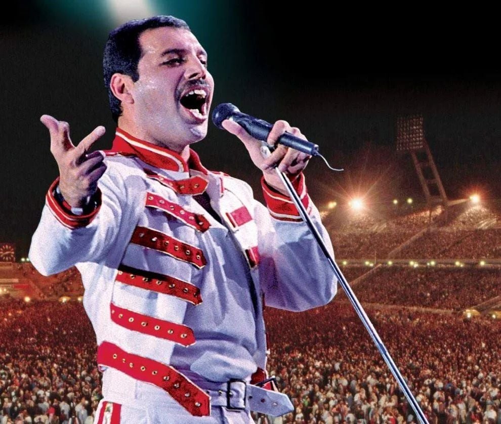Sänger in weiß-rotem Kostüm singt auf einer großen Bühne vor einem vollen Stadion.