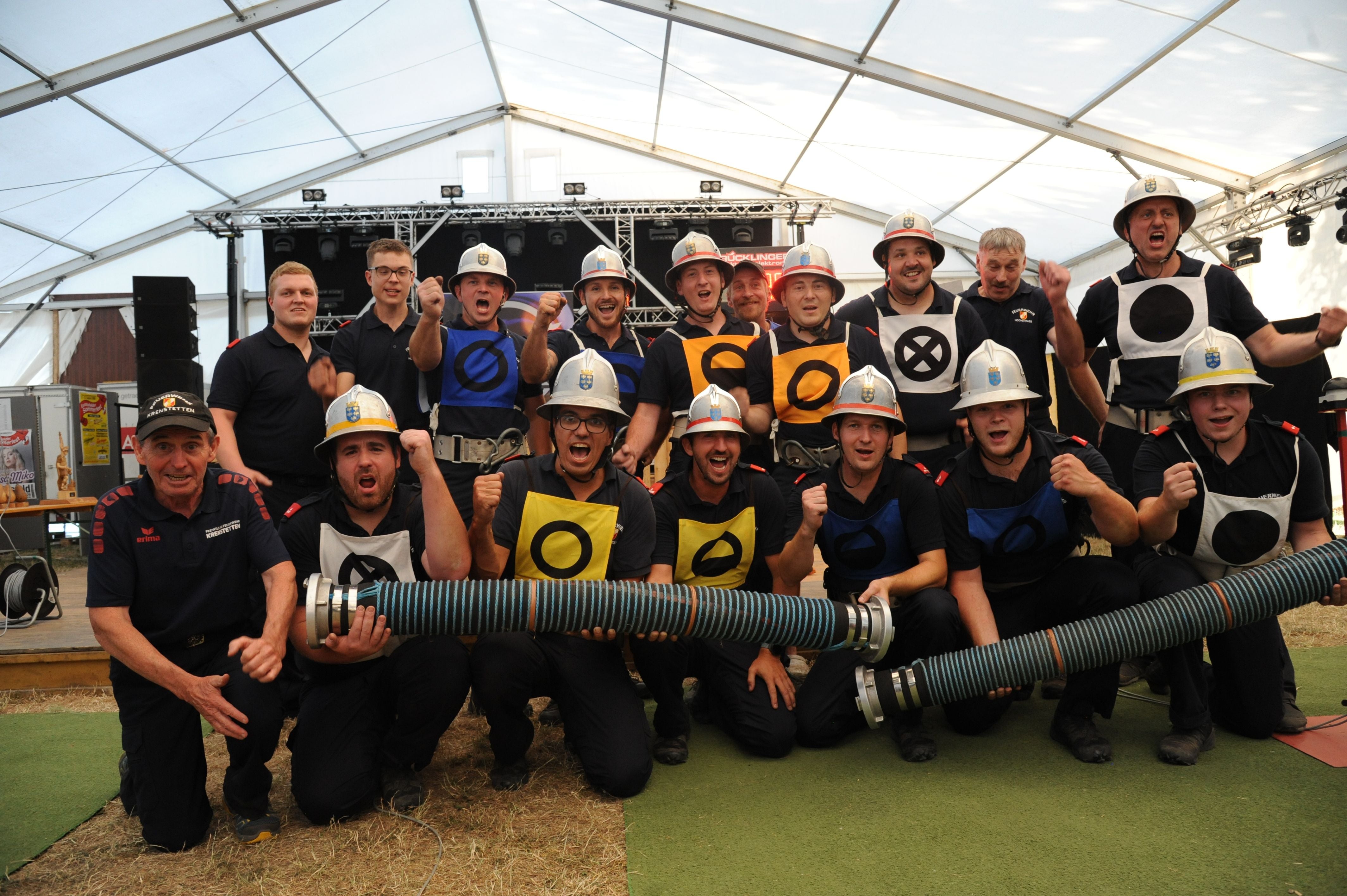 Eine Gruppe Feuerwehrleute posiert jubelnd mit Helmen und farbigen Markierungswesten im Festzelt.