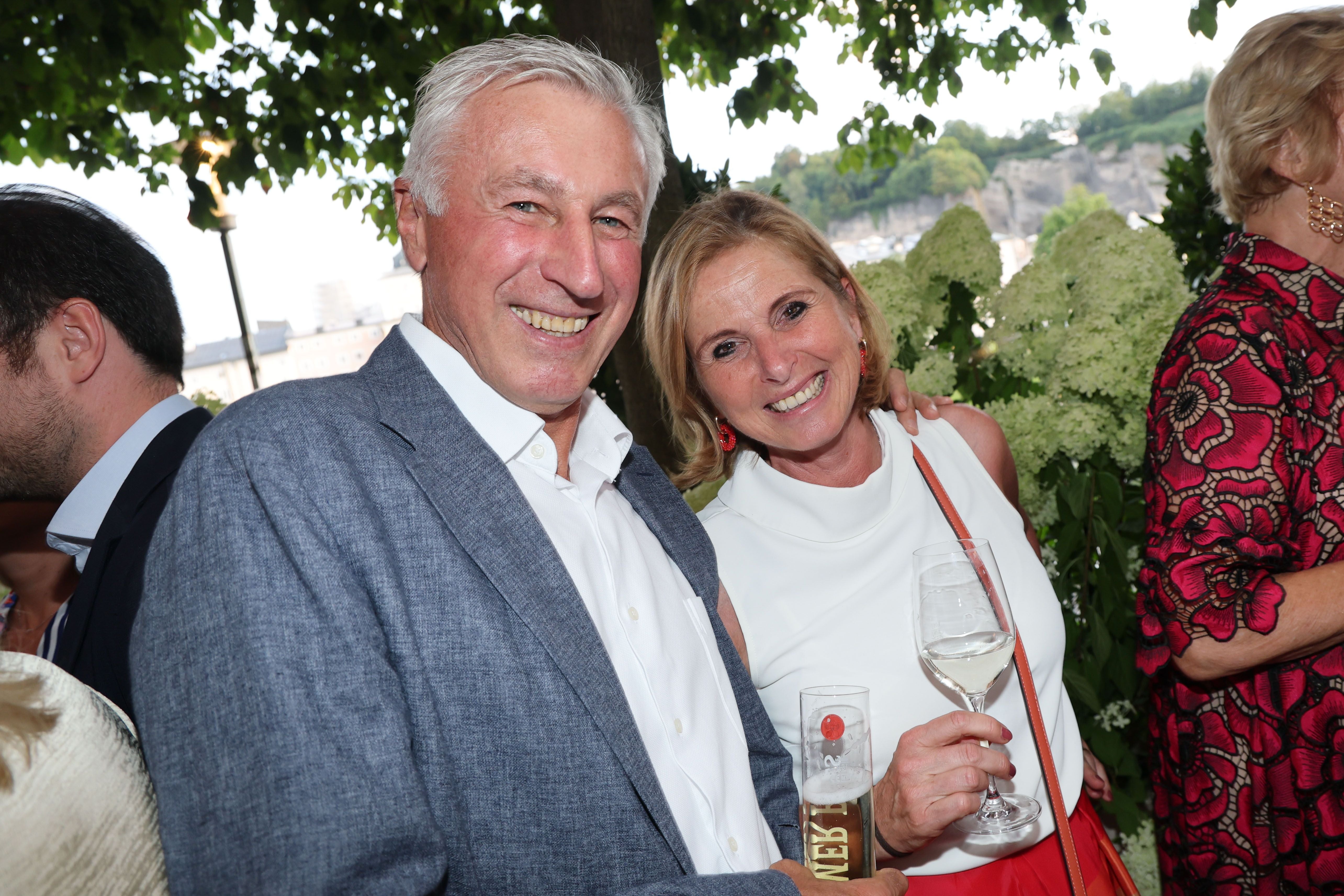 Ein älteres Paar lächelt auf einer Gartenparty und hält jeweils ein Glas mit Getränk.