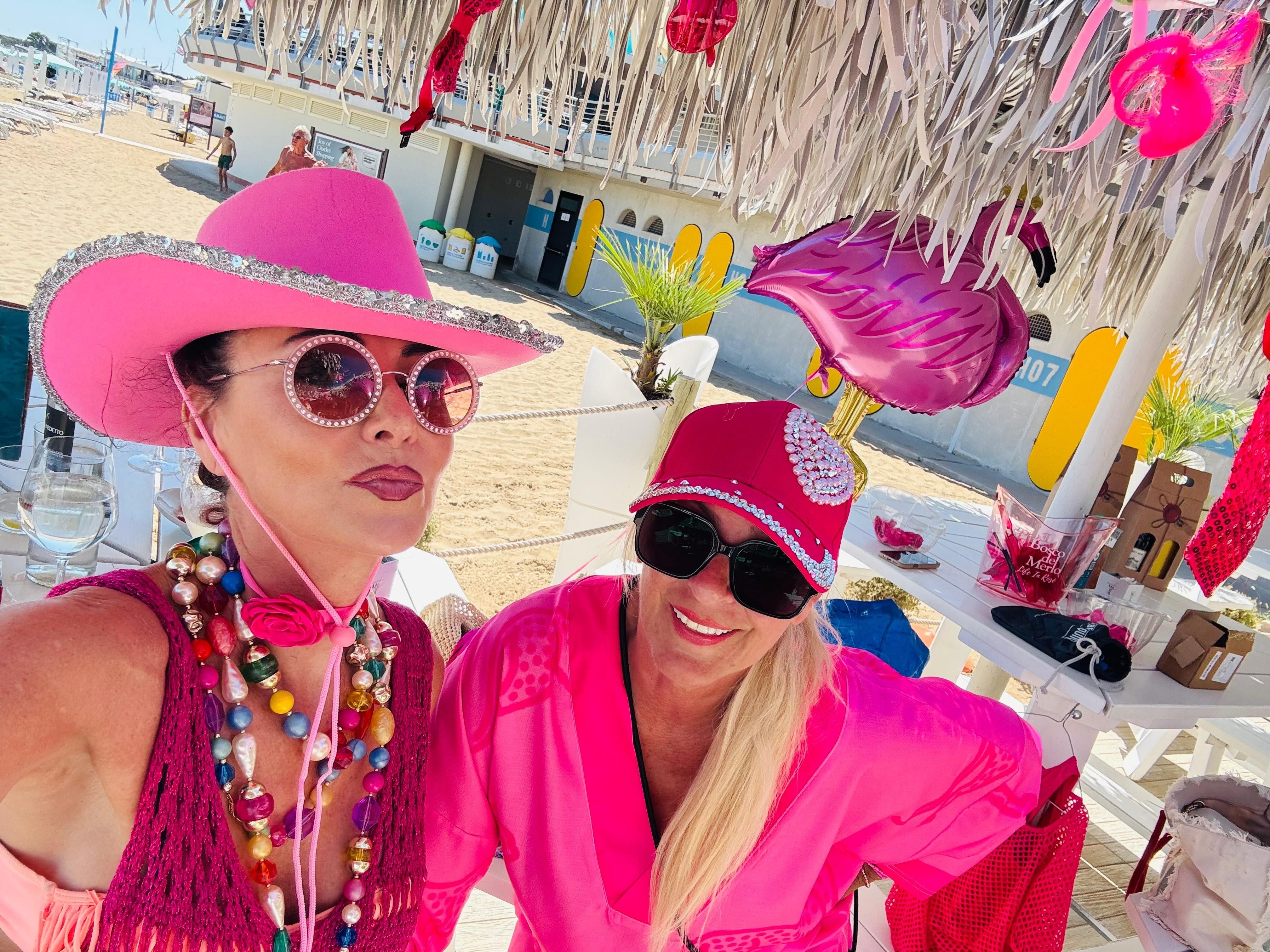 Zwei Frauen in pinker Kleidung und Sonnenbrillen am Strand unter einer dekorierten Hütte.