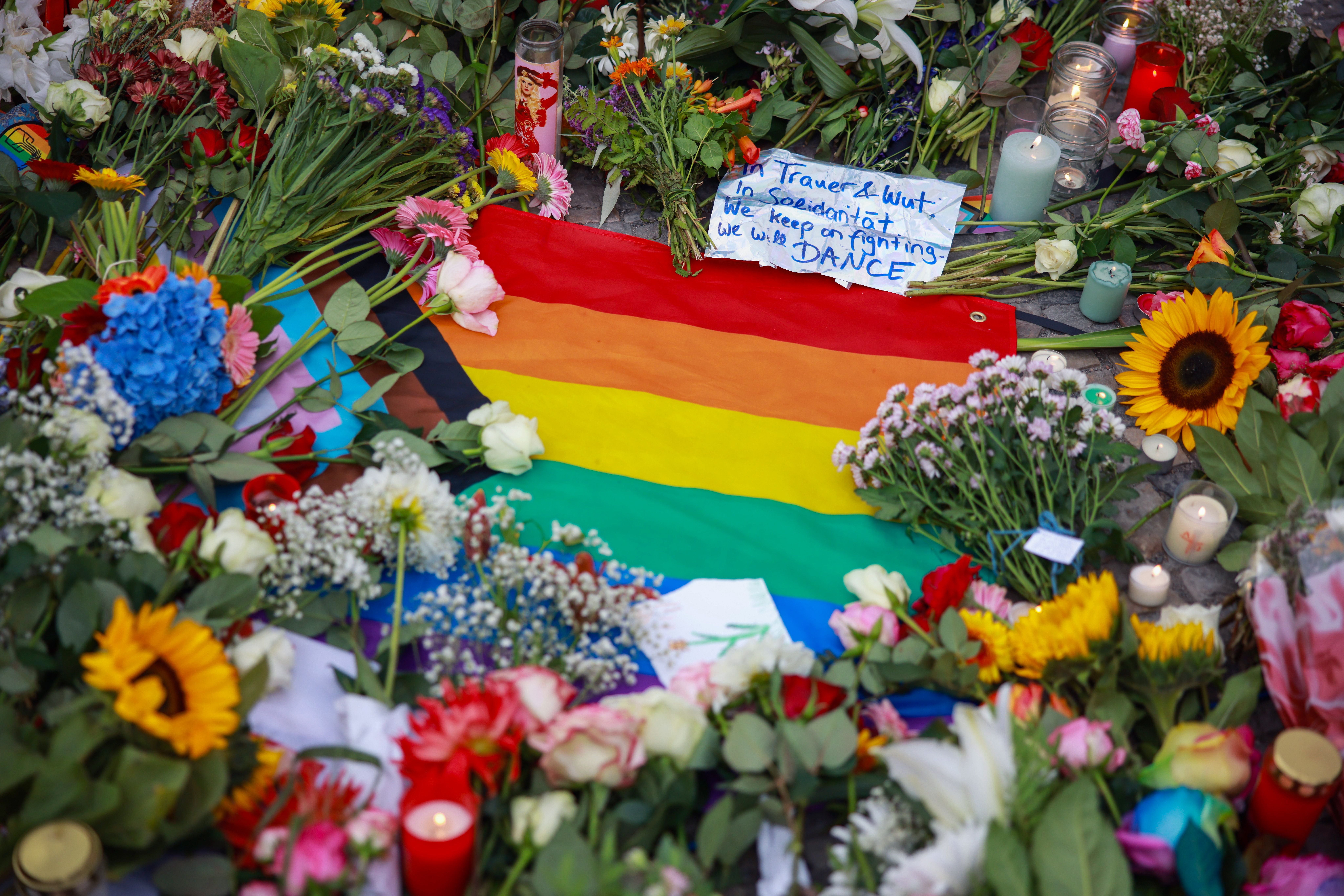 Regenbogenflagge, Blumen, Kerzen und ein Trauerzettel bilden ein Gedenkarrangement auf dem Boden.