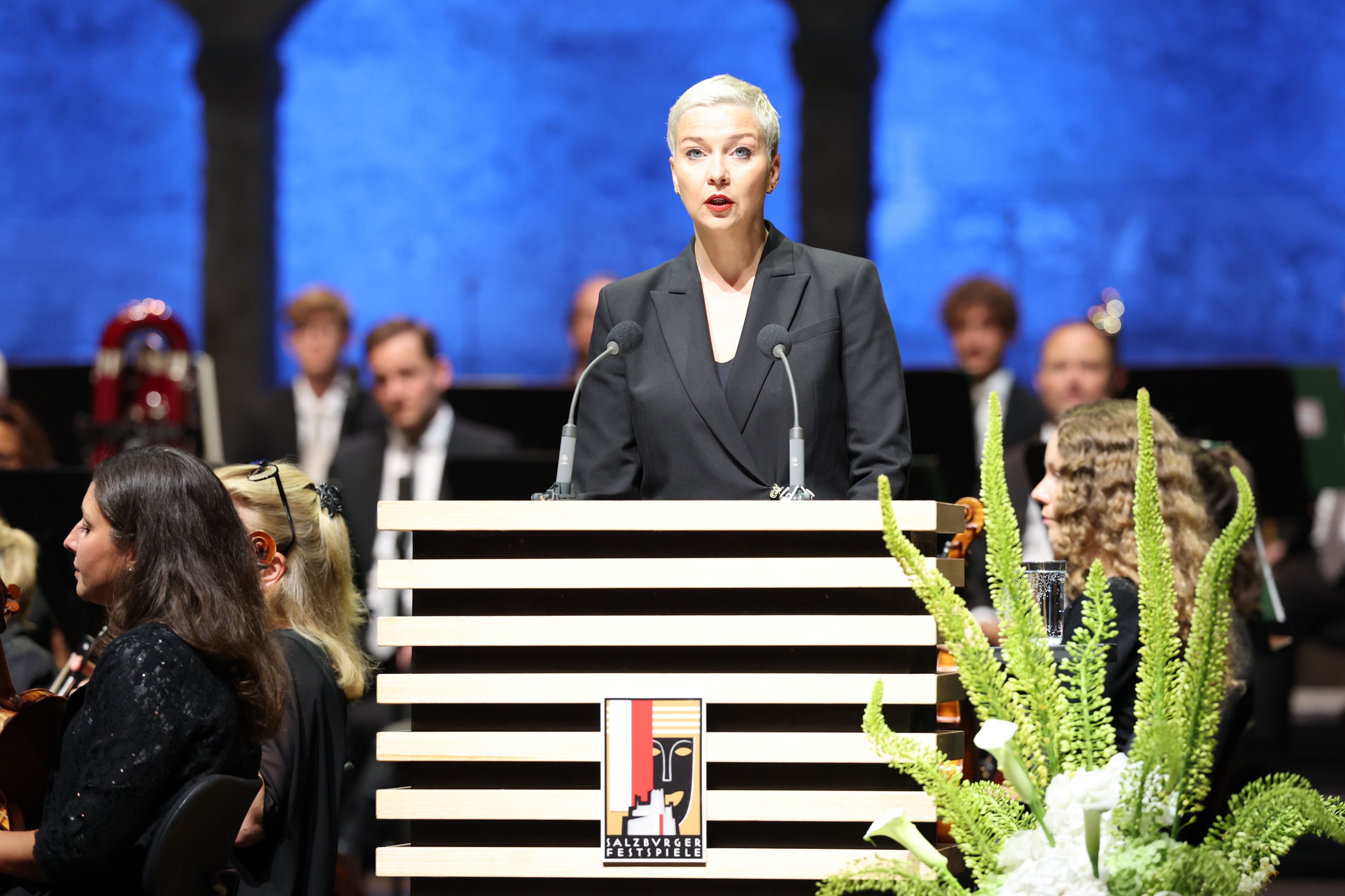 Eine Frau hält eine Eröffnungsrede bei den Salzburger Festspielen, Orchester im Hintergrund.