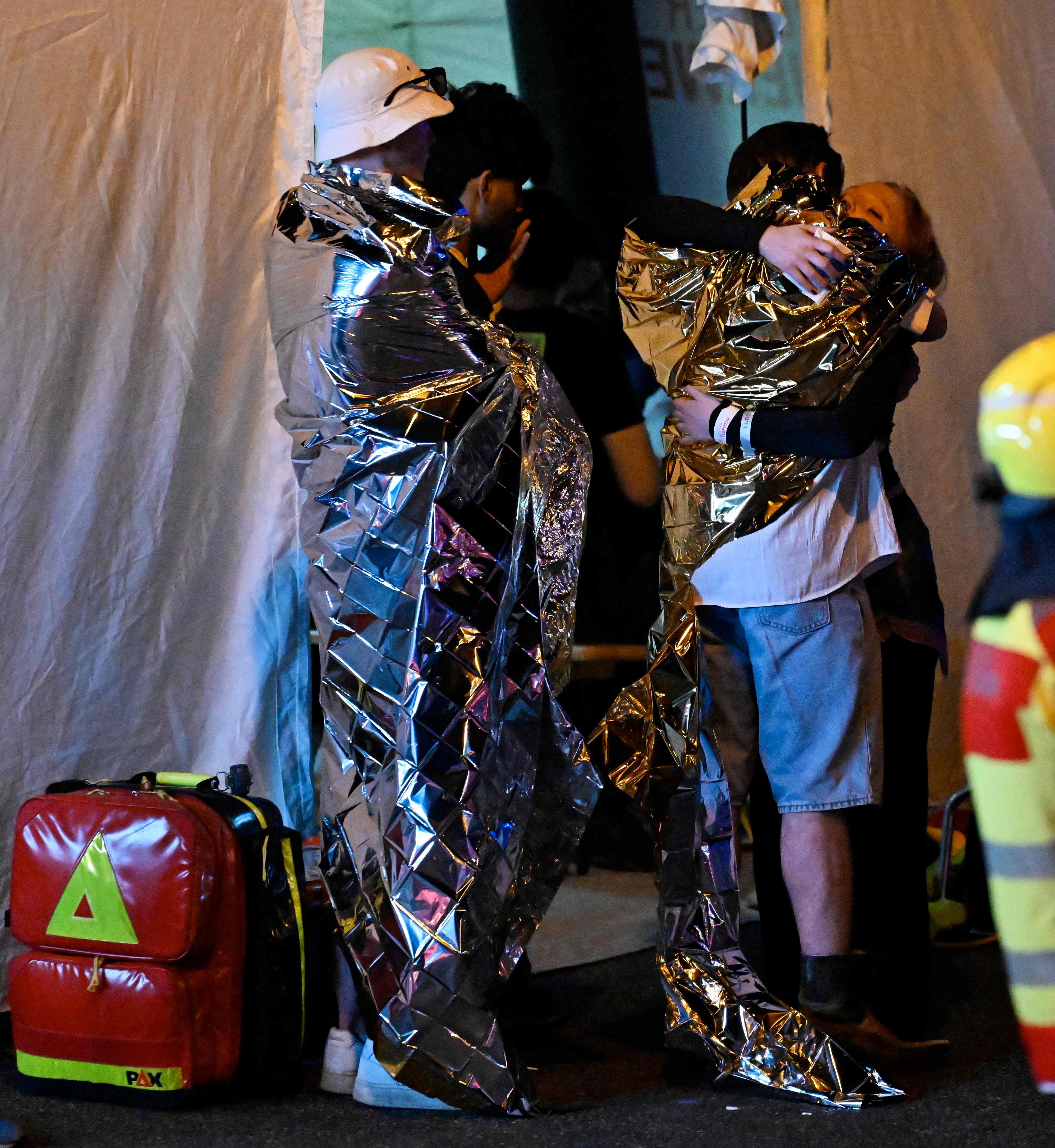 Menschen in Rettungsdecken umarmen sich neben einem Notfallrucksack bei Nacht.