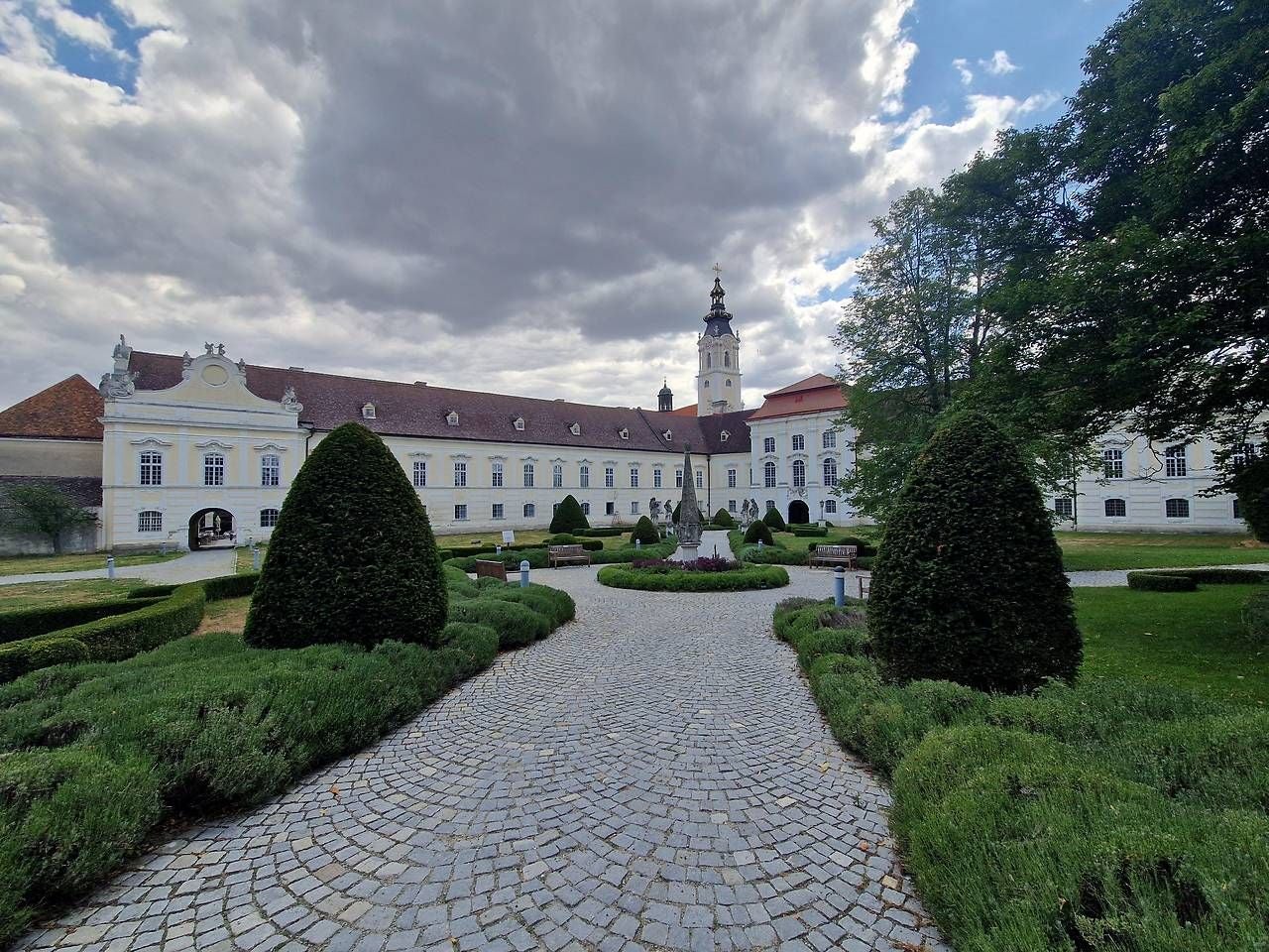 Barockes Kloster Altenburg mit gepflegtem Garten, Hecken und wolkigem Himmel im Hintergrund.