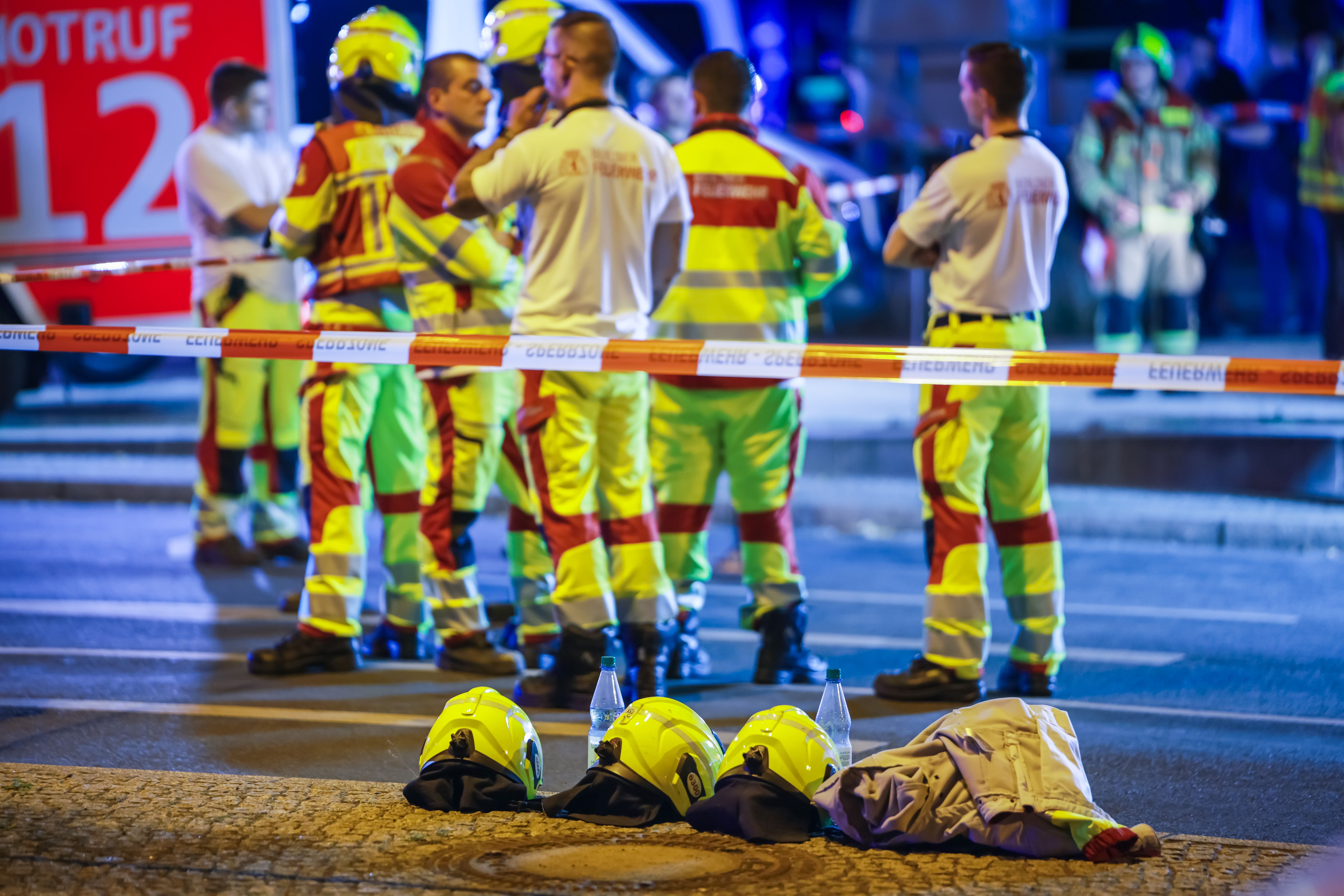 Mehrere Rettungskräfte stehen hinter Absperrband, Helme und Ausrüstung liegen im Vordergrund.