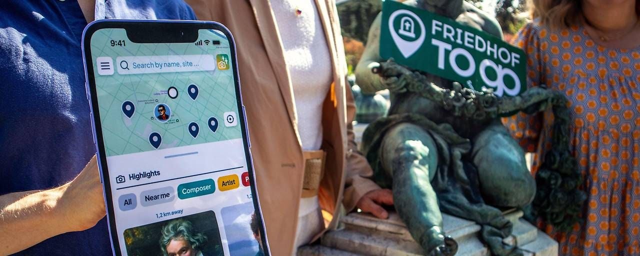Drei Personen zeigen eine Handy-App vor einer Statue am Wiener Zentralfriedhof.