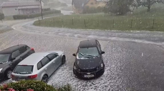 Starker Hagel bedeckt die Straße und geparkte Autos in einer ländlichen Gegend in der Steiermark.