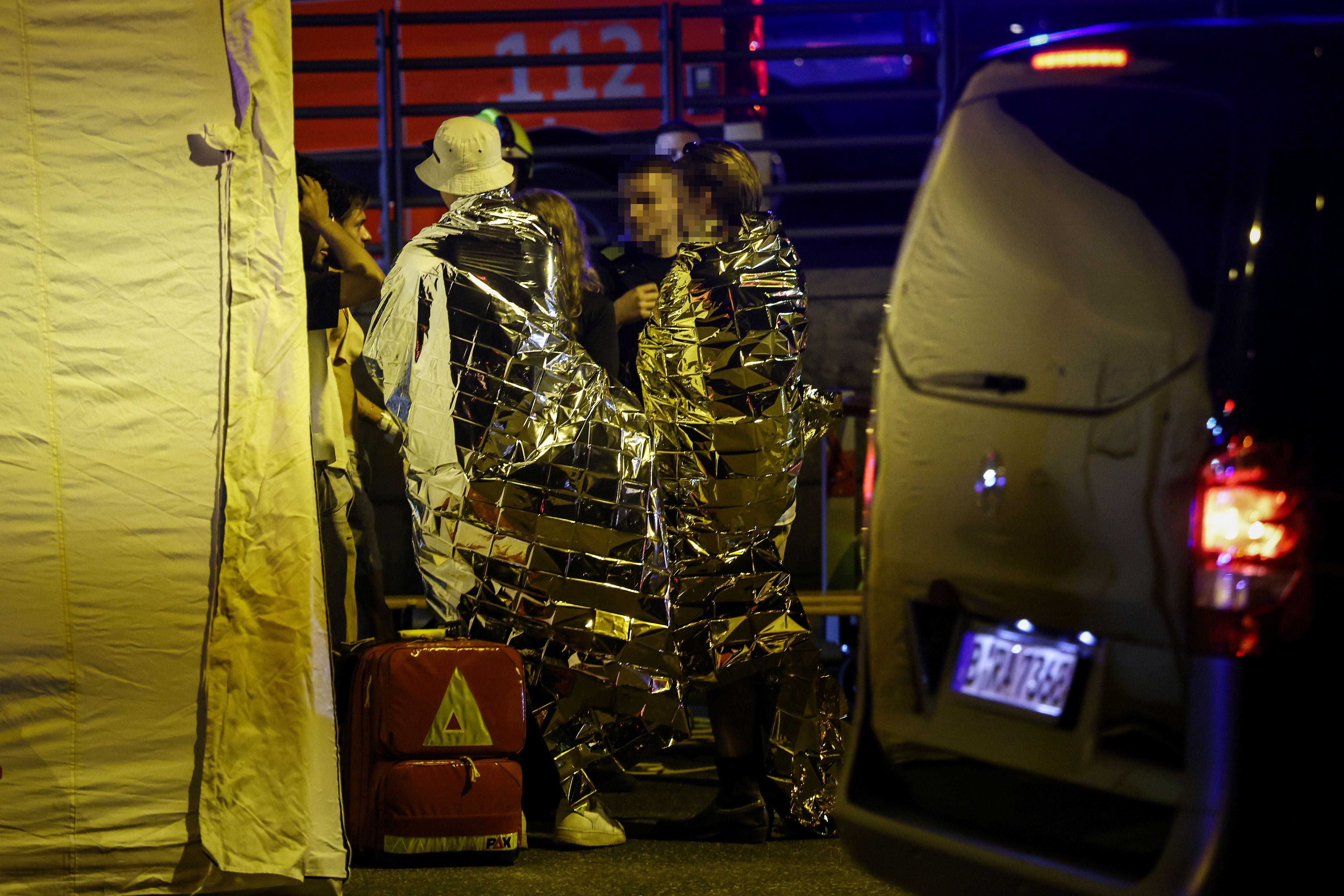 Menschen in Rettungsdecken stehen an einem Notfallzelt neben einem Krankentransportwagen bei Nacht.