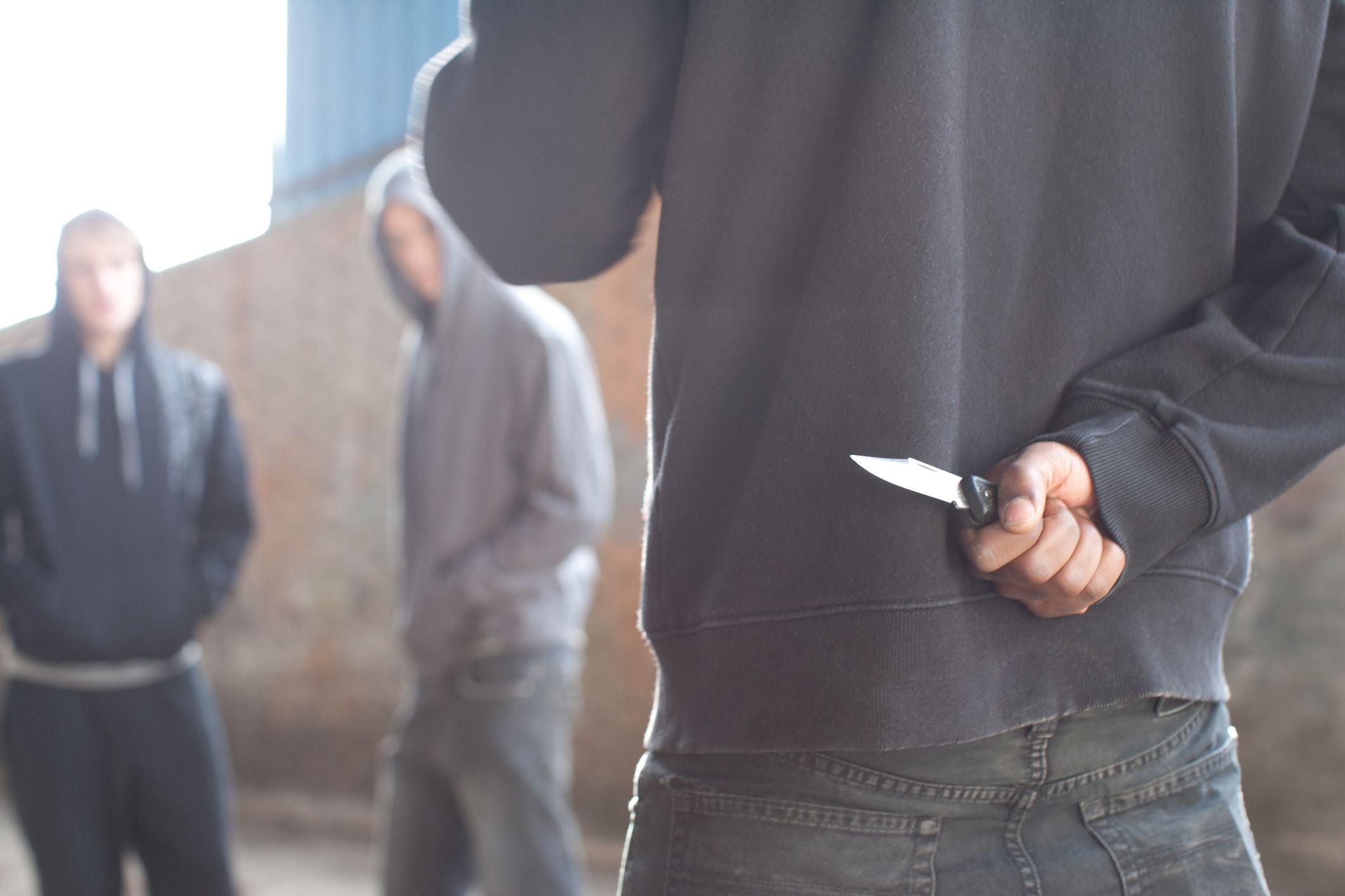 Eine Person hält ein Messer hinter dem Rücken, zwei weitere Personen stehen im Hintergrund.