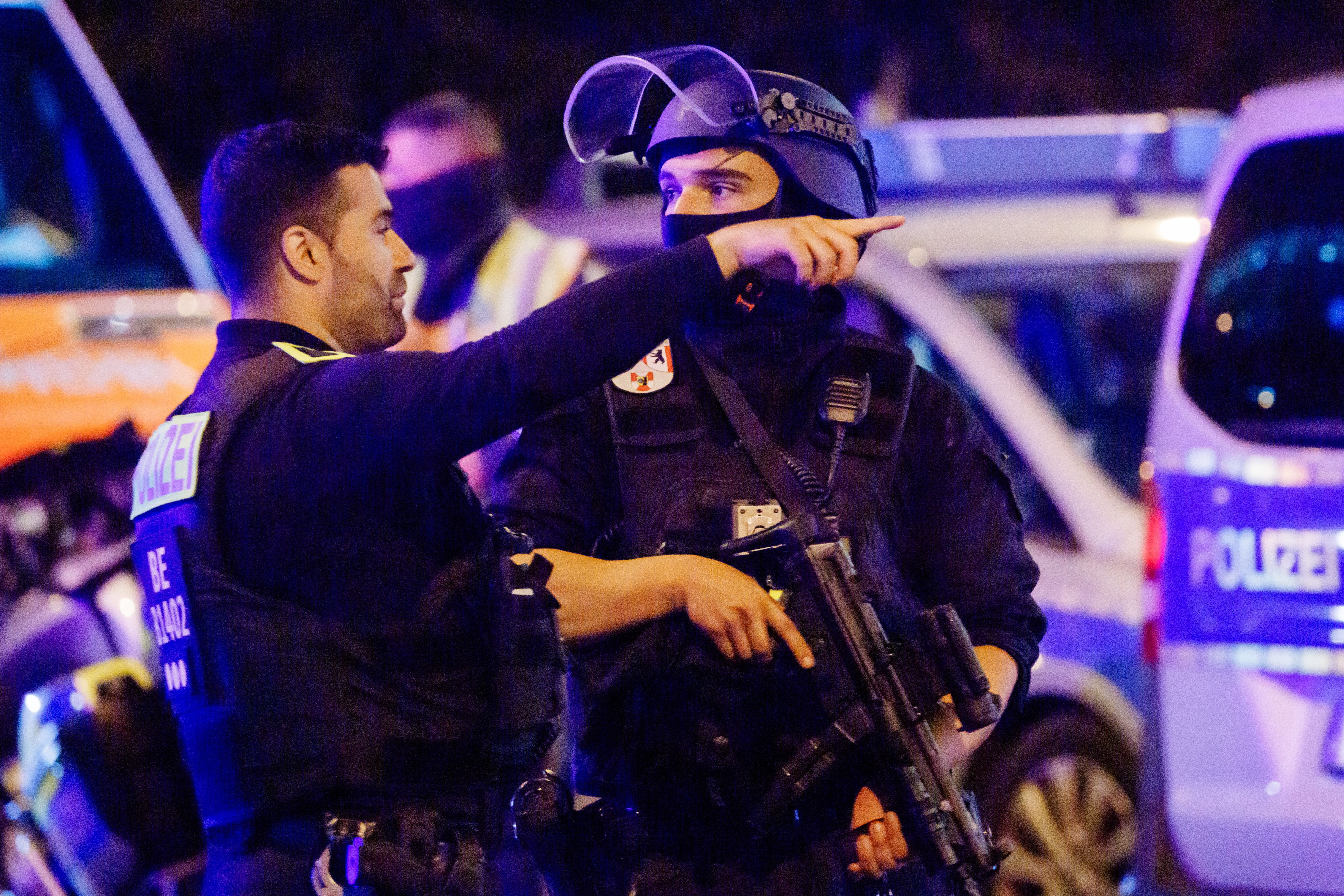 Zwei Polizisten reden, einer davon hält ein Maschinengewehr, im Hintergrund Polizeiautos.