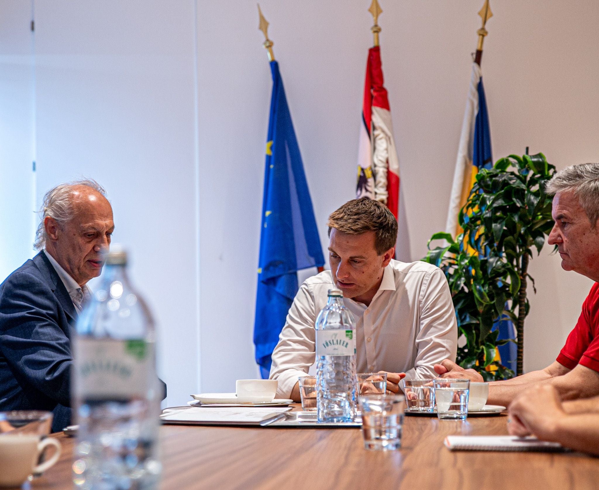 Drei Männer sitzen an einem Konferenztisch mit Wasserflaschen und Flaggen im Hintergrund.