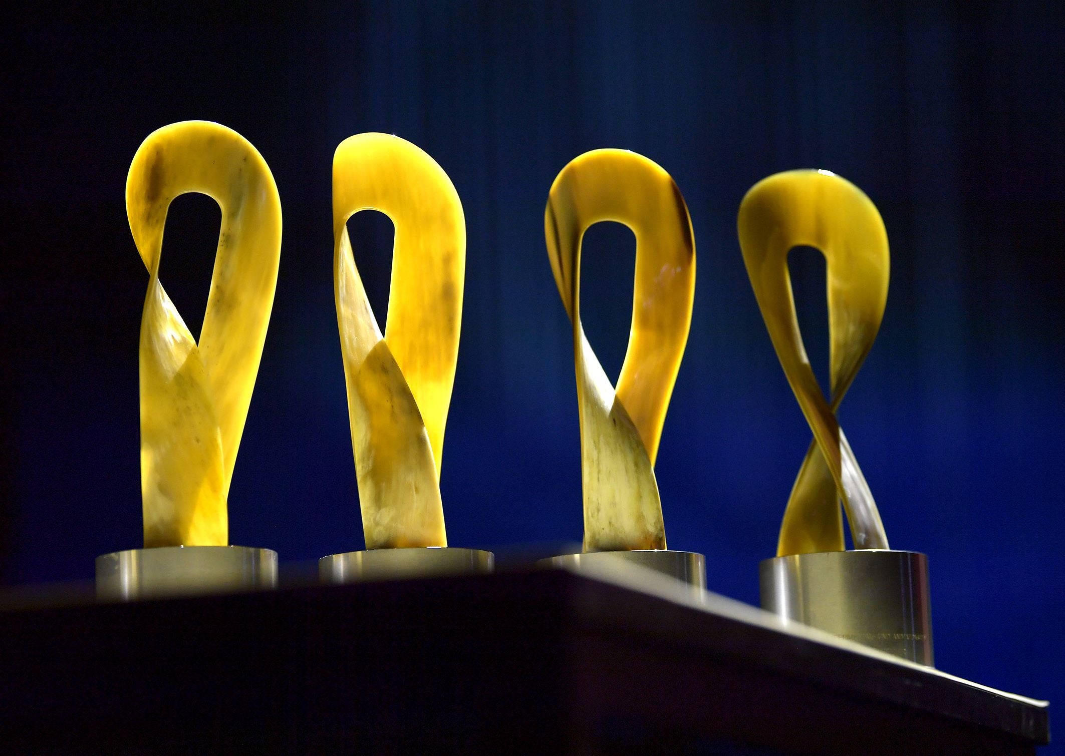 Vier goldene Trophäen mit geschwungenem Design stehen nebeneinander auf einem Tisch.