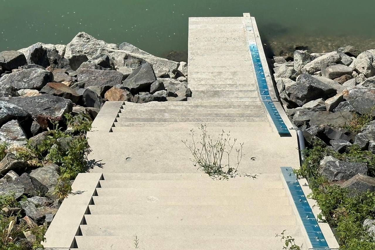 Betontreppe mit Wasserstandsmesslatte führt zu einem Fluss, umgeben von Felsen und Pflanzen.