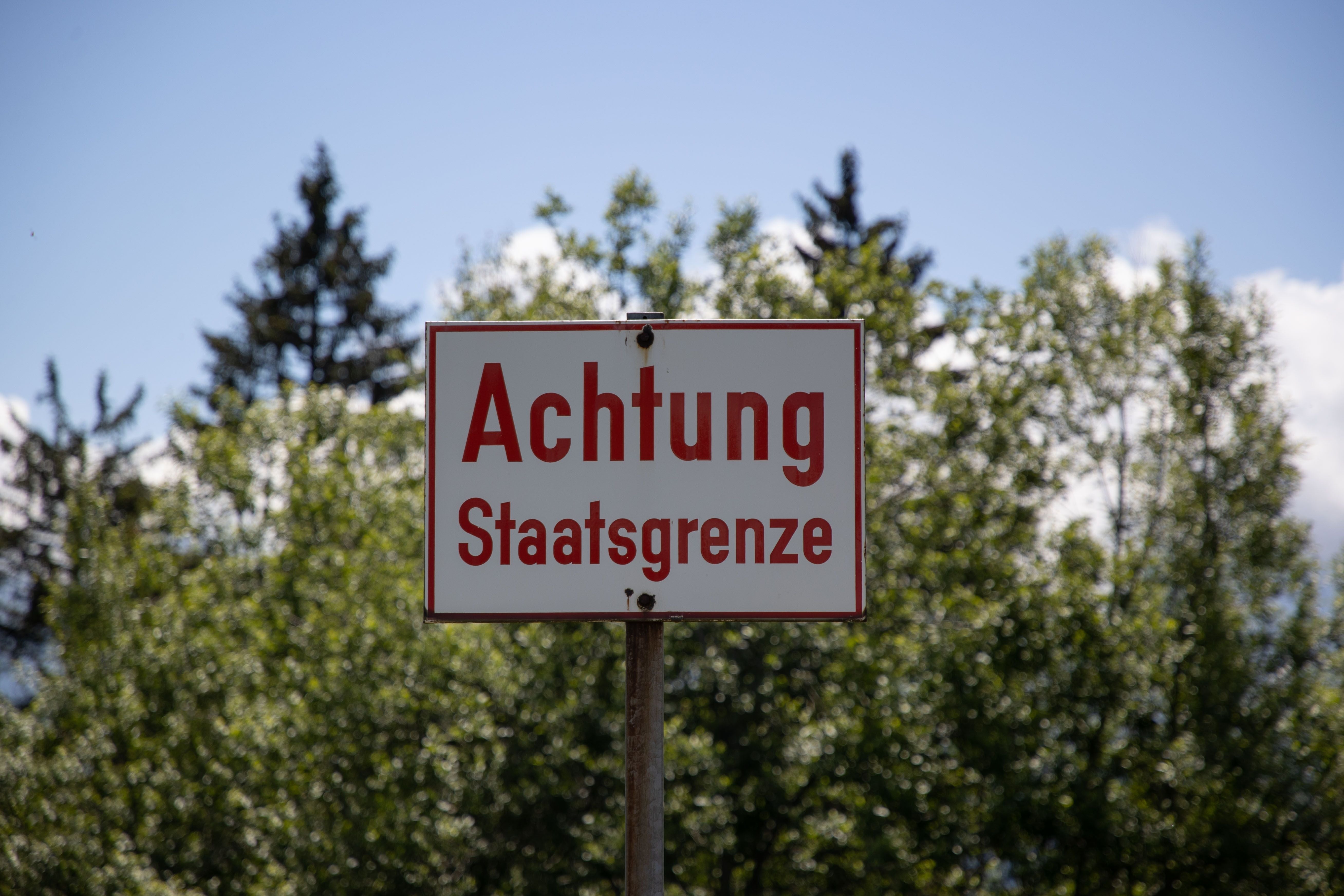 Hinweisschild mit der Aufschrift „Achtung Staatsgrenze“ vor Bäumen im Hintergrund.