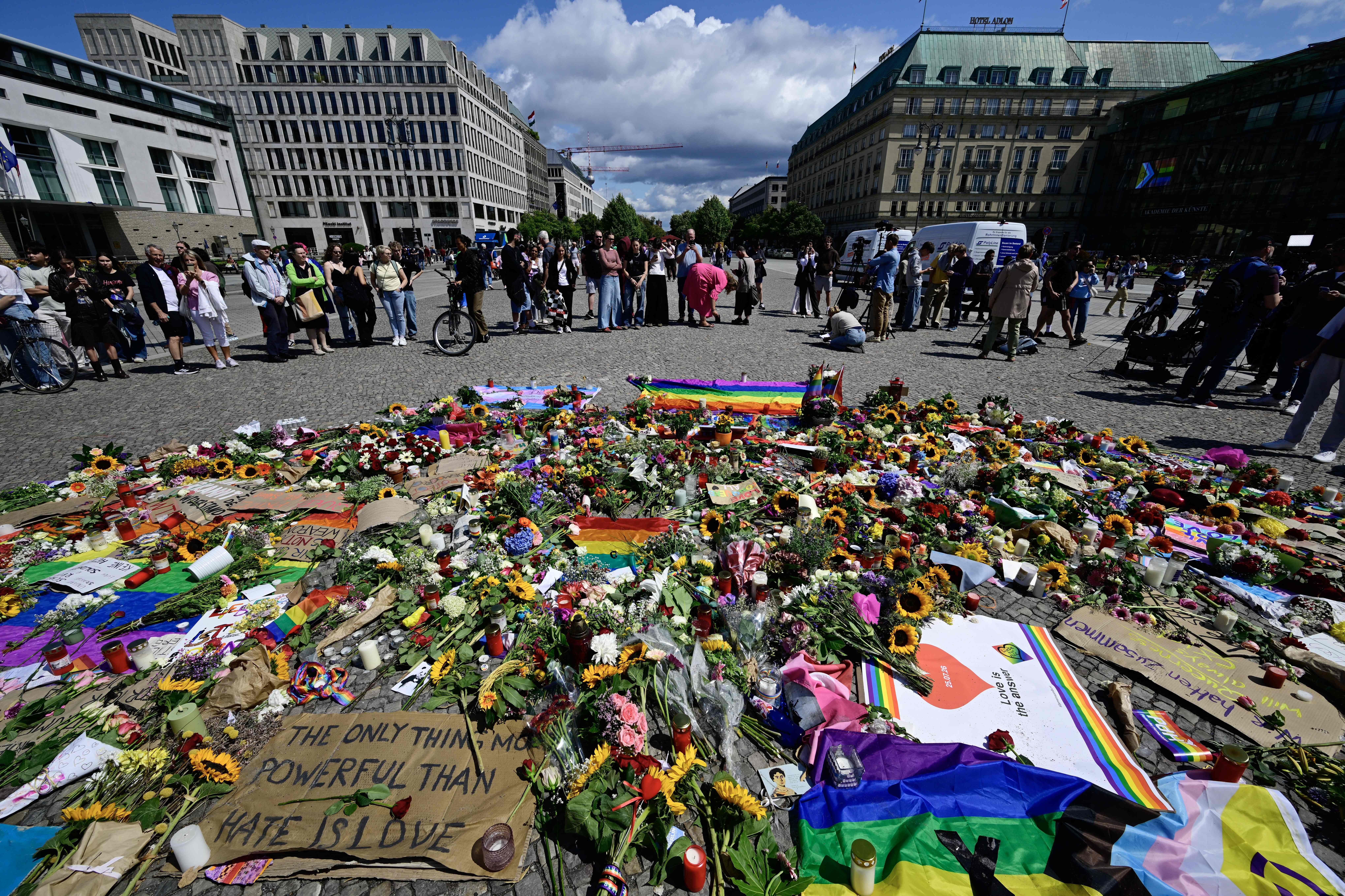 Menschen stehen vor Blumen, Kerzen, Regenbogenflaggen und Trauerplakaten am Pariser Platz in Berlin.