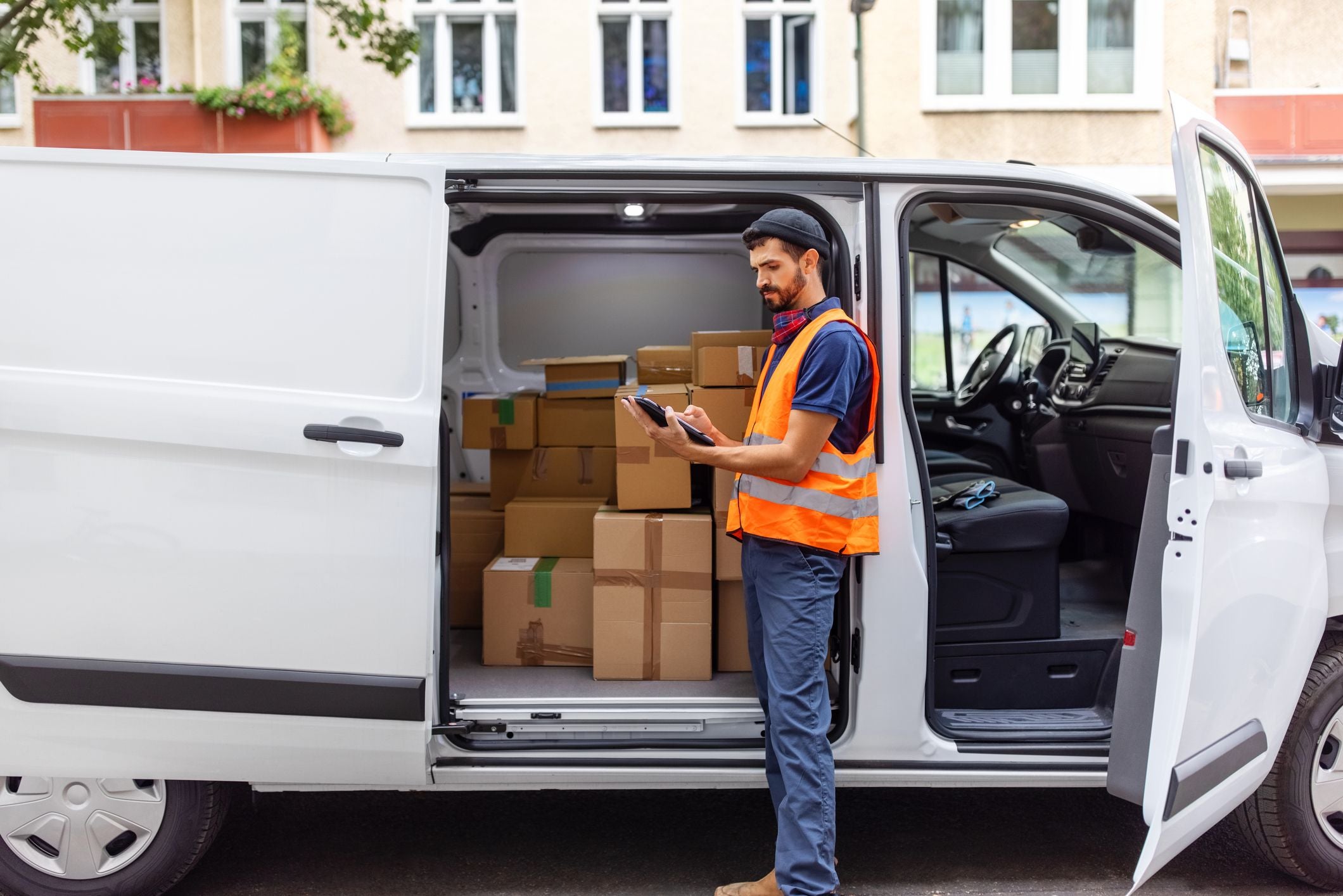 Paketzusteller in Warnweste prüft Lieferungen am Tablet neben offenem Lieferwagen voller Pakete.
