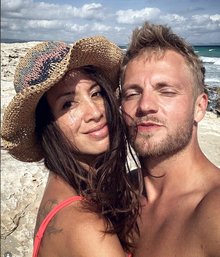 Junges Paar macht ein Selfie am Strand, beide lächeln, sie trägt einen Strohhut.