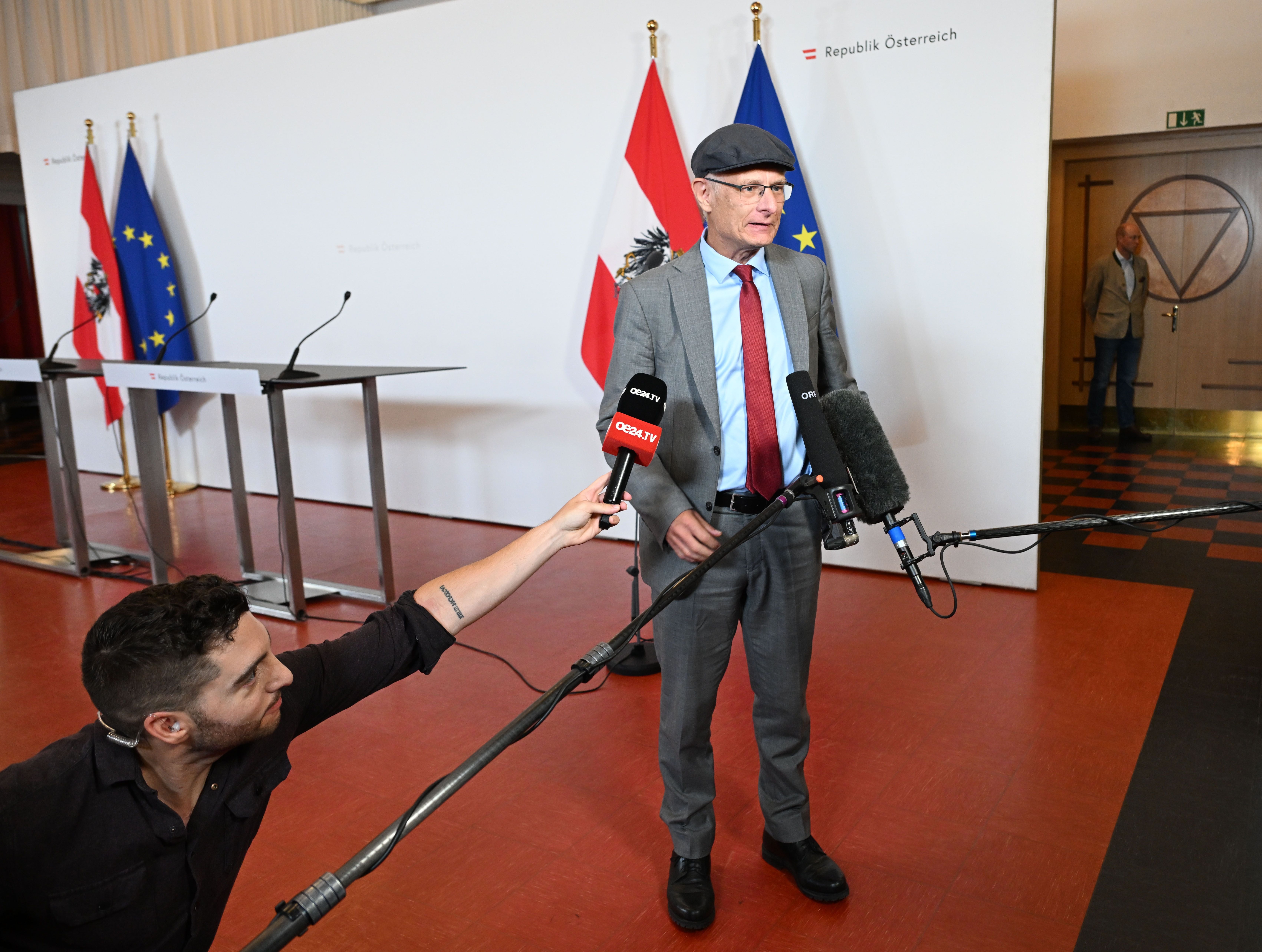 Ein Mann im Anzug spricht vor Mikrofonen bei einer Pressekonferenz mit Fahnen im Hintergrund.