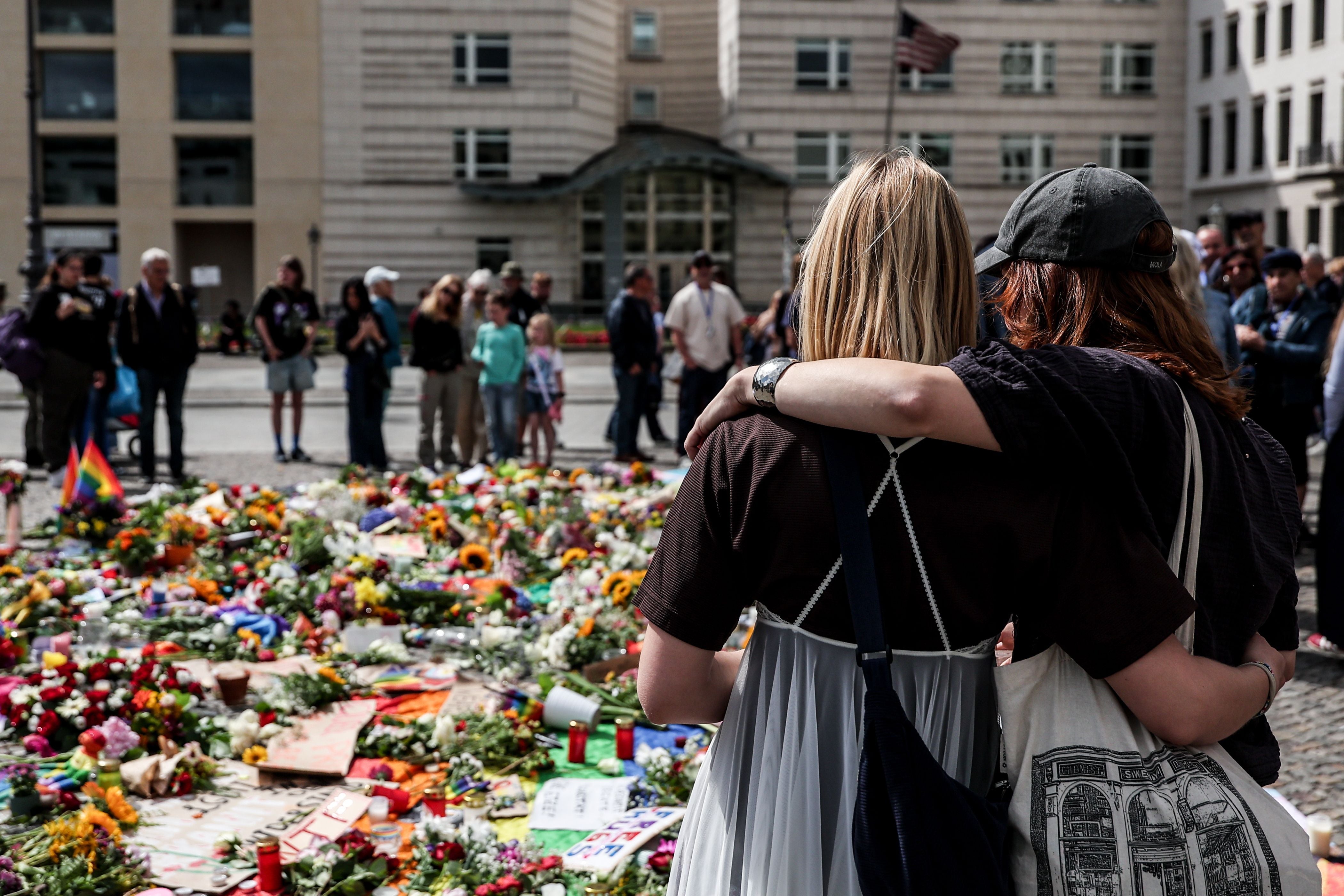 Zwei trauernde Personen umarmen sich vor Blumen und Kerzen an einer Gedenkstätte in Berlin.