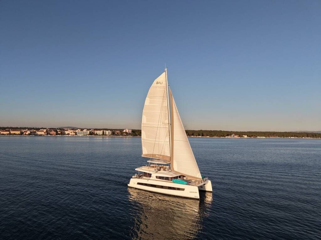 Ein weißer Katamaran segelt auf ruhigem Wasser vor einer Küstenstadt bei klarem Himmel.
