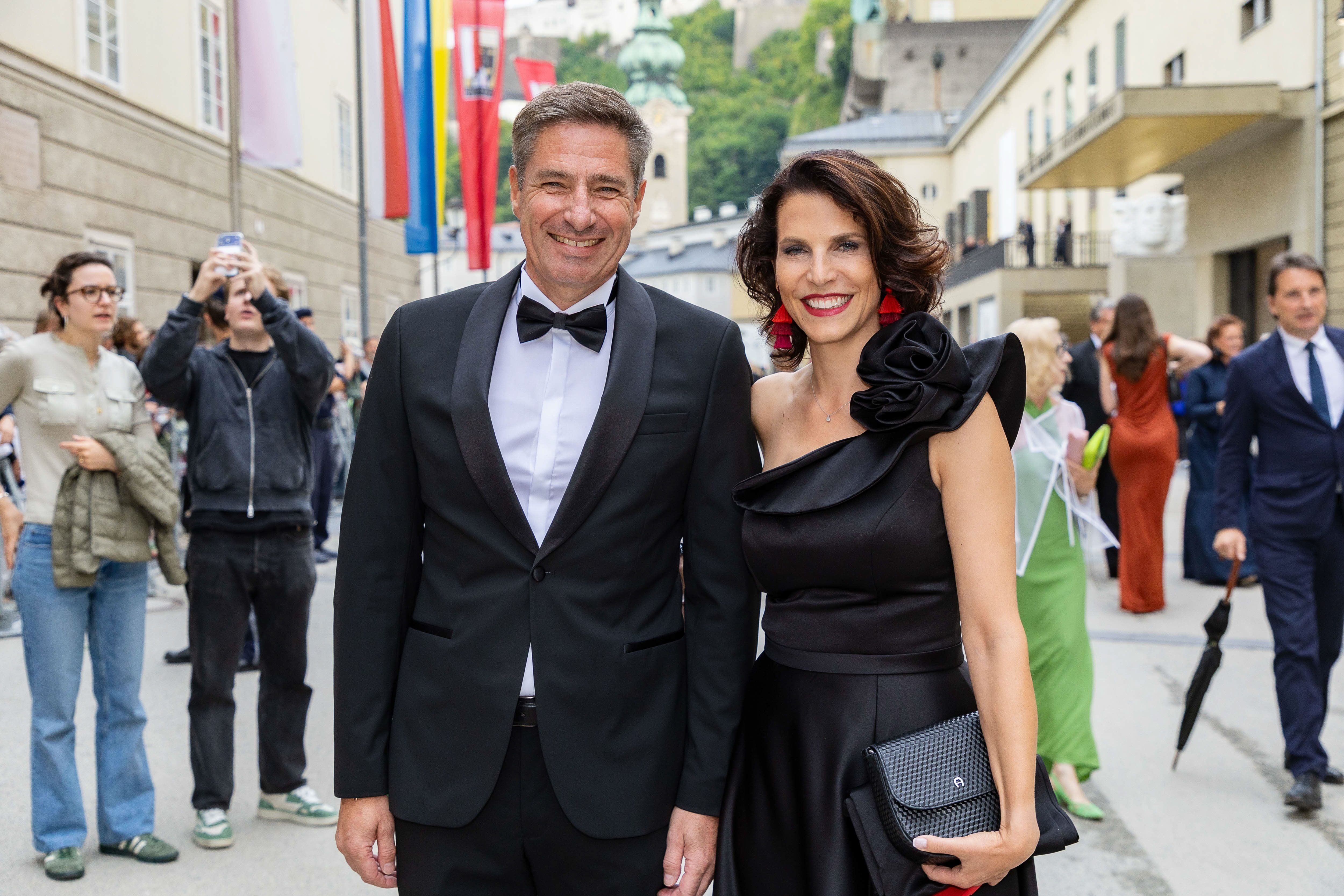 Ein Mann im Smoking und eine Frau im schwarzen Kleid lächeln bei einer Premiere im Freien.