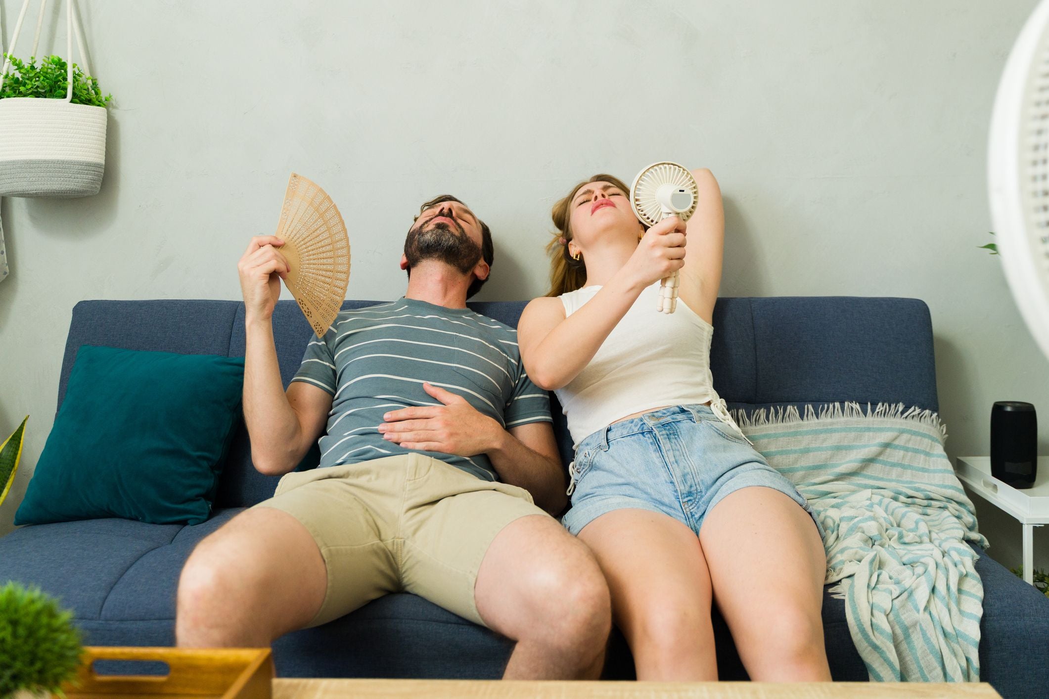 Ein erschöpftes Paar sitzt schwitzend auf dem Sofa und kühlt sich mit Fächern und Ventilator ab.