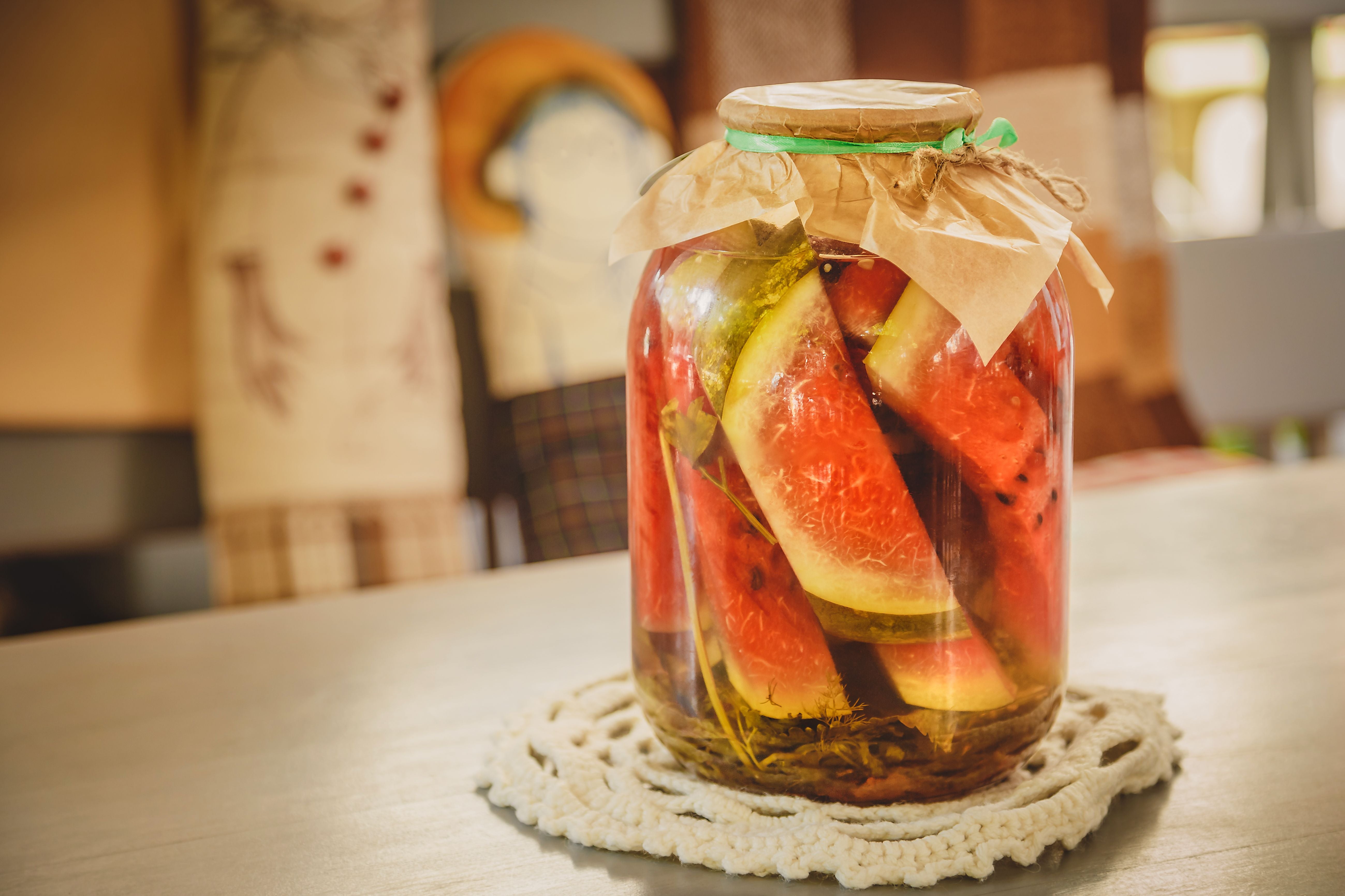 Ein Glas mit eingelegter Wassermelone und Kräutern steht auf einem Holz­tisch in einer Küche.