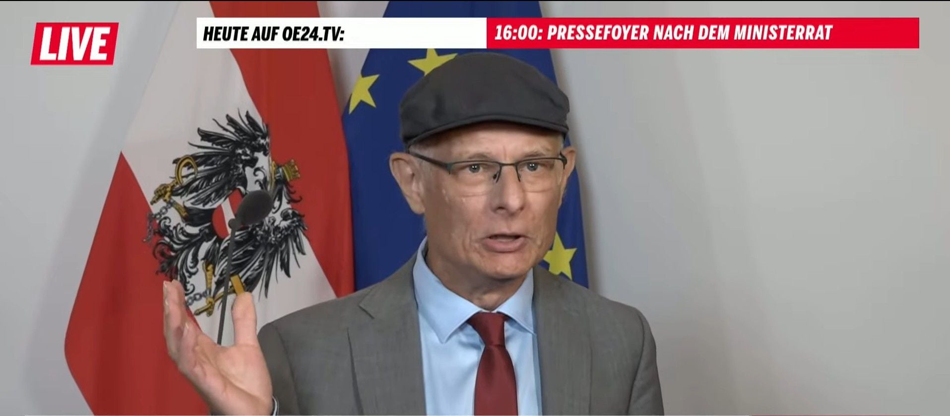 Älterer Mann mit Brille und Mütze spricht vor österreichischer und EU-Flagge auf einer Pressekonferenz.