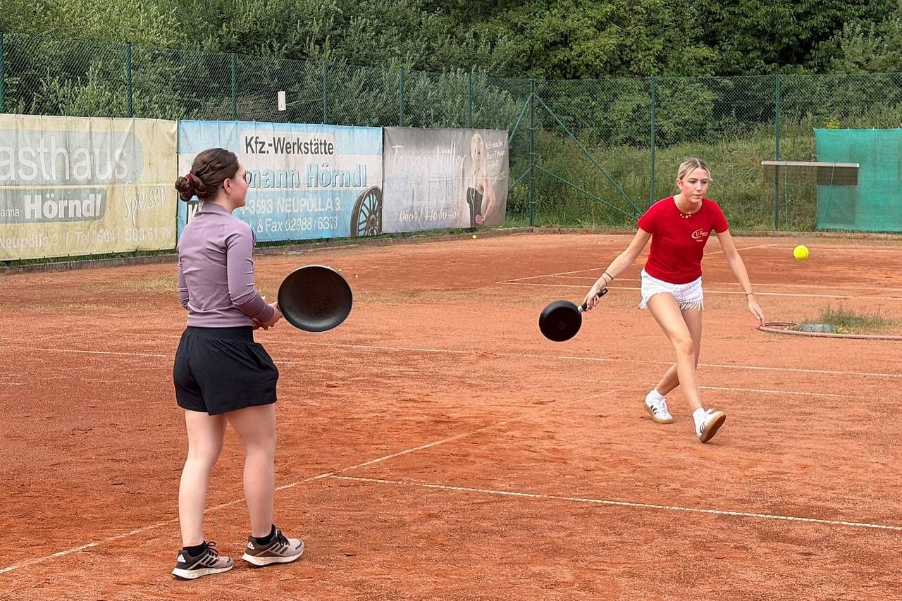 Zwei junge Frauen spielen Tennis auf Sandplatz, benutzen dabei jedoch Bratpfannen statt Tennisschläger.