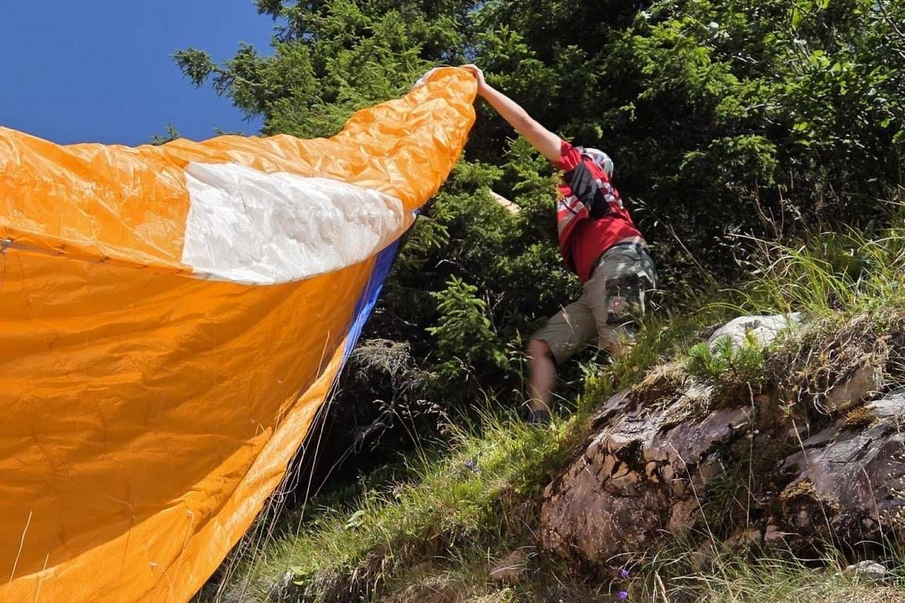 Person mit orange-weißem Gleitschirm hängt im Hang neben Felsen und Bäumen fest.