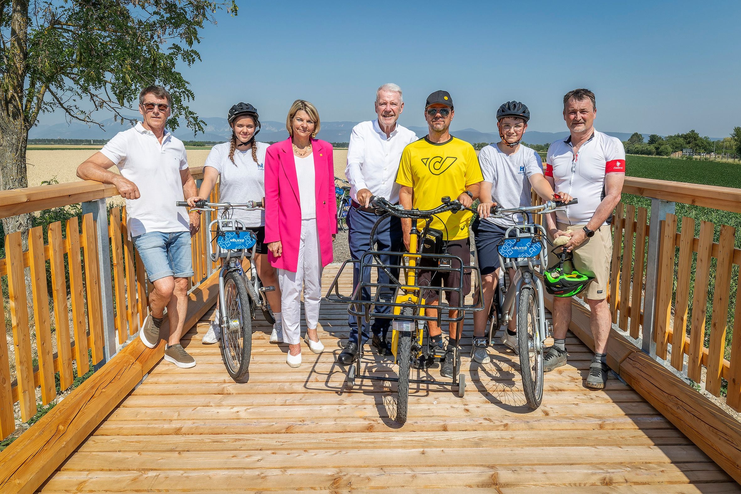 Sieben Personen stehen mit Fahrrädern auf einer Holzbrücke bei sonnigem Wetter in der Natur.