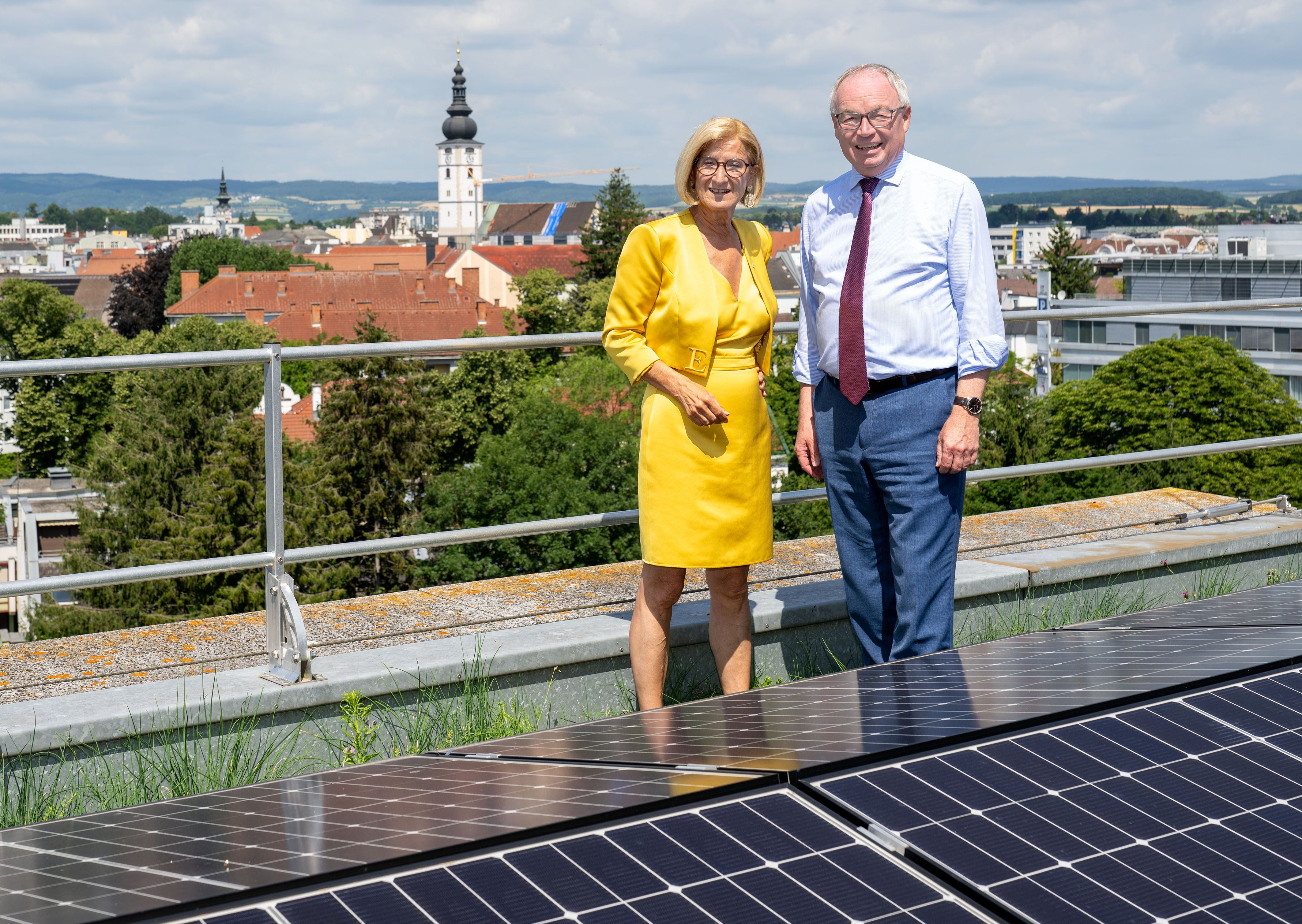 Eine Frau und ein Mann stehen neben Solarpaneelen auf einem Dach mit Stadtpanorama im Hintergrund.