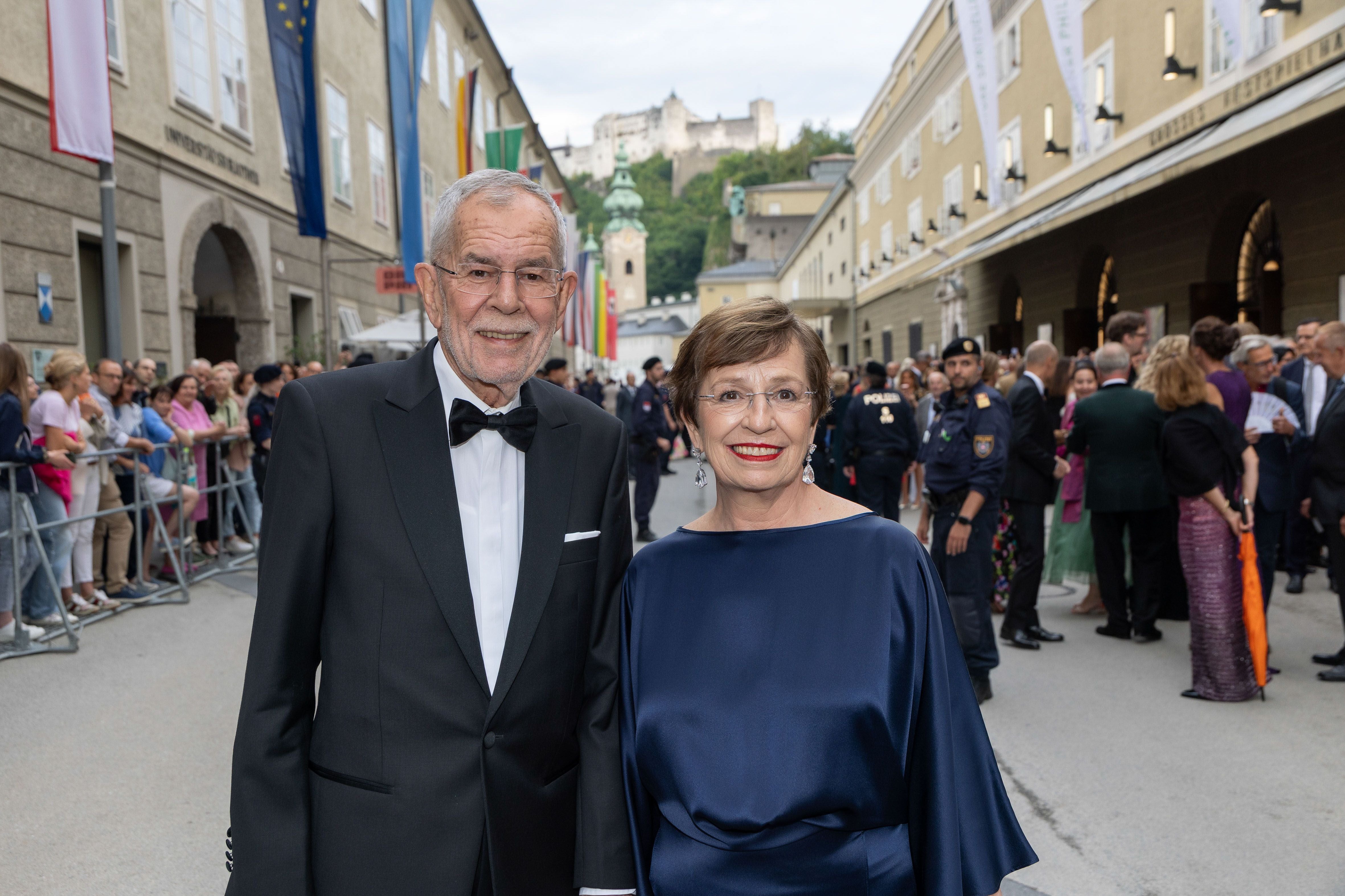 Ein elegant gekleidetes Paar steht auf einer Festspielstraße mit Menschenmenge im Hintergrund.