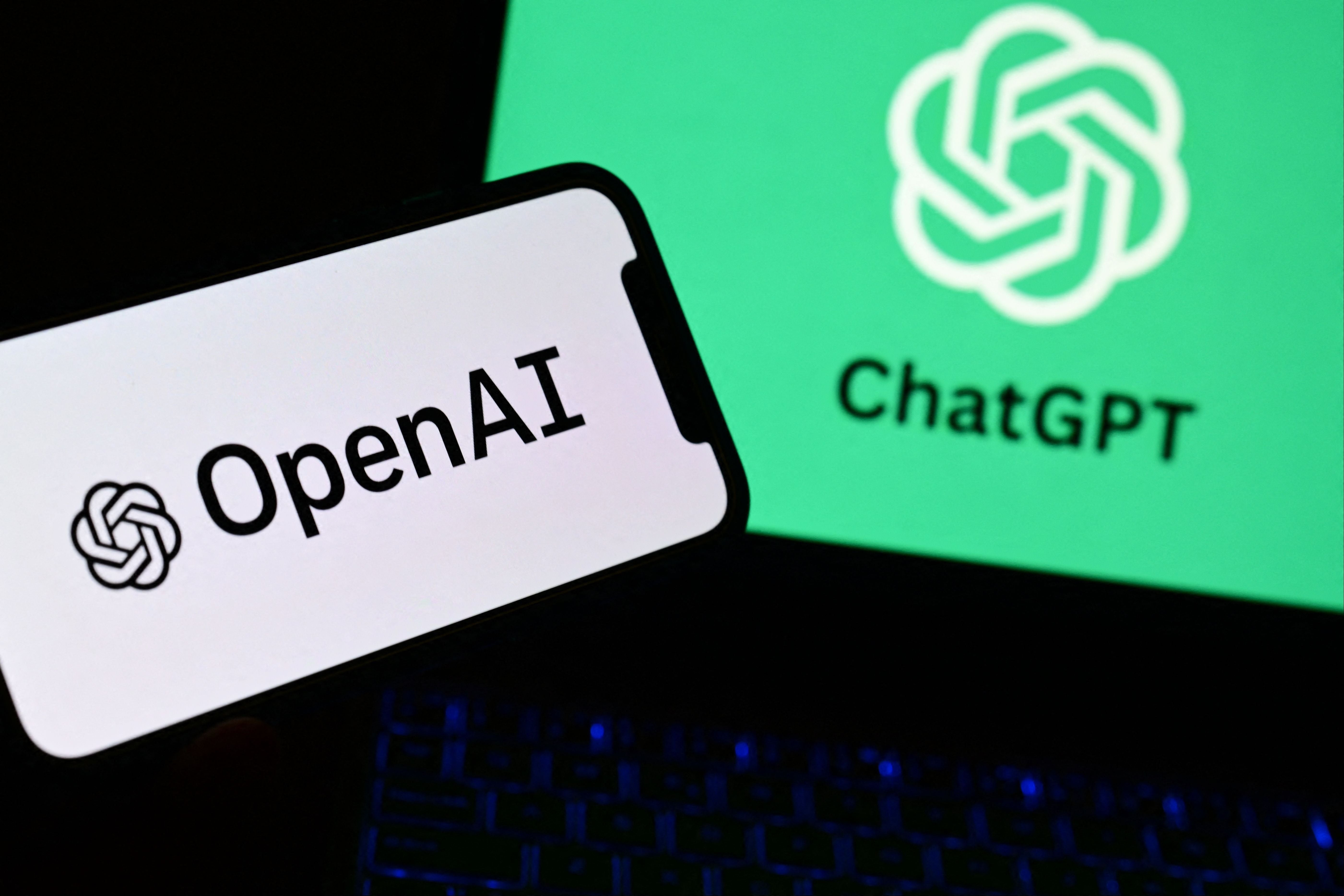 Ein Smartphone zeigt das OpenAI-Logo, ein Laptop im Hintergrund das ChatGPT-Logo.