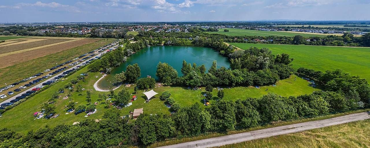 Luftaufnahme des Badeteichs von Laxenburg mit umliegenden Wiesen und vielen parkenden Autos.