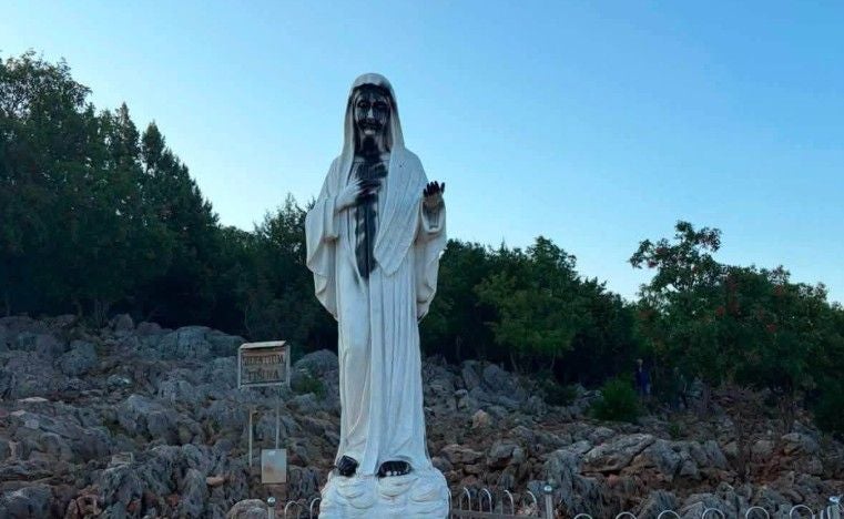 Beschädigte Marienstatue steht auf steinigem Boden mit dunklen Flecken und Bäumen im Hintergrund.