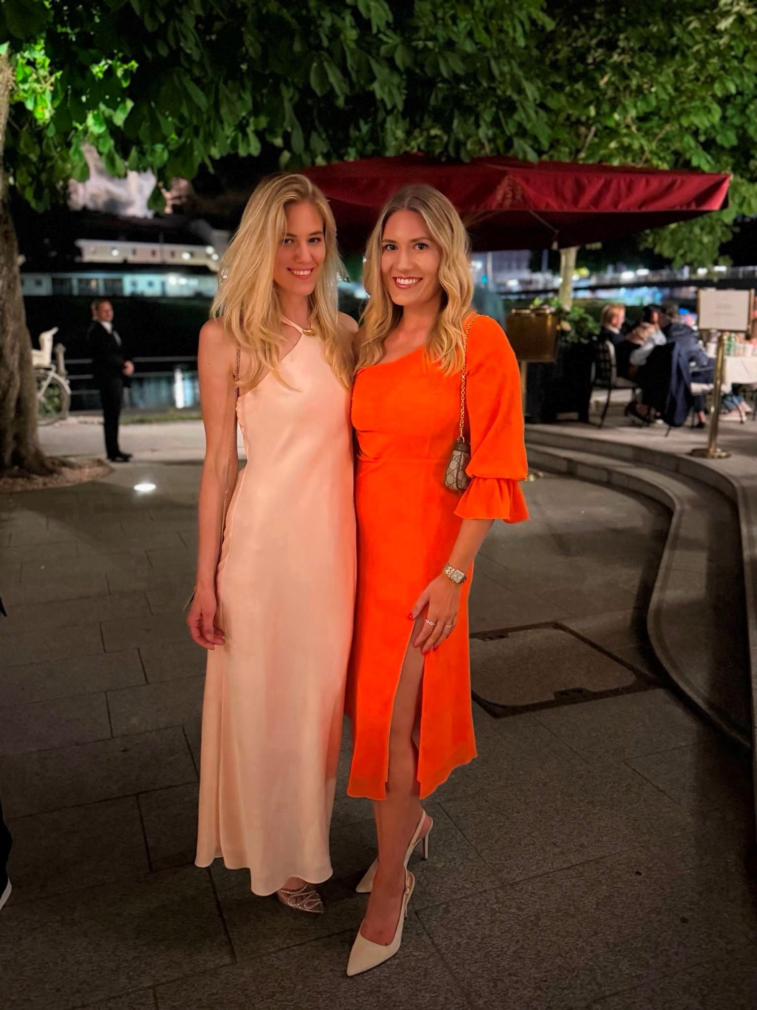 Zwei Frauen in eleganten Kleidern posieren abends draußen vor einem Restaurant.