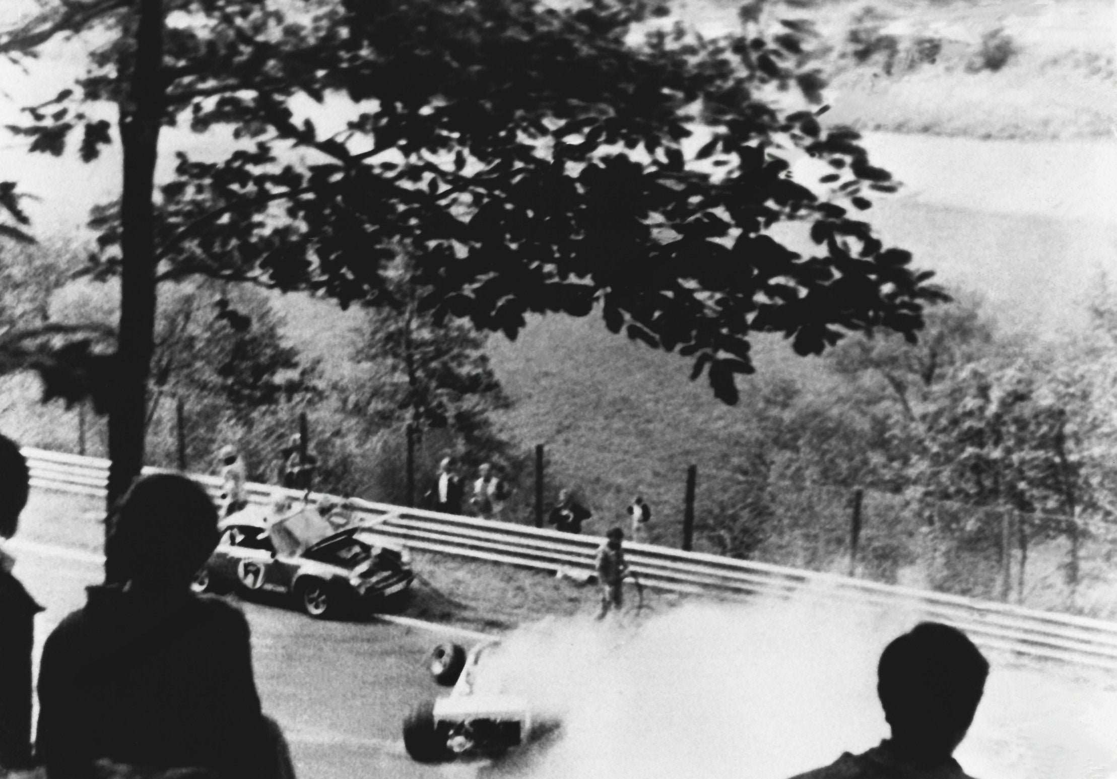 Schwarzweißfoto eines Autounfalls auf einer Rennstrecke mit qualmendem Rennwagen und Zuschauern im Vordergrund.