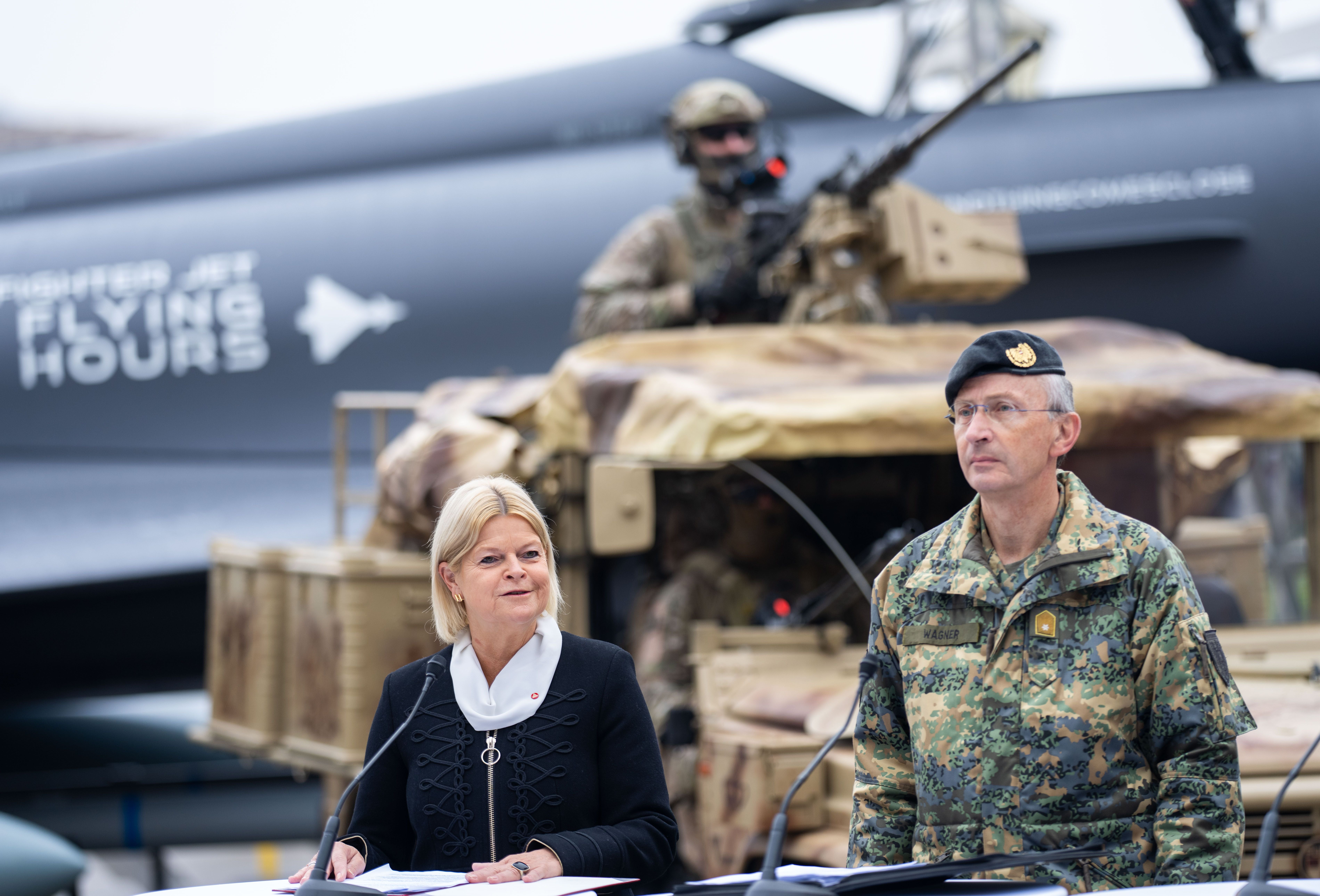 Verteidigungsministerin und Offizier sprechen bei einer Pressekonferenz vor Militärfahrzeugen.