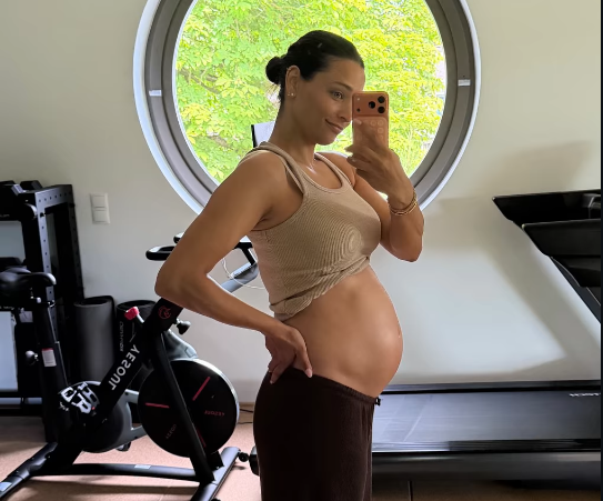 Schwangere Frau macht ein Selfie im Fitnessraum vor einem runden Fenster.