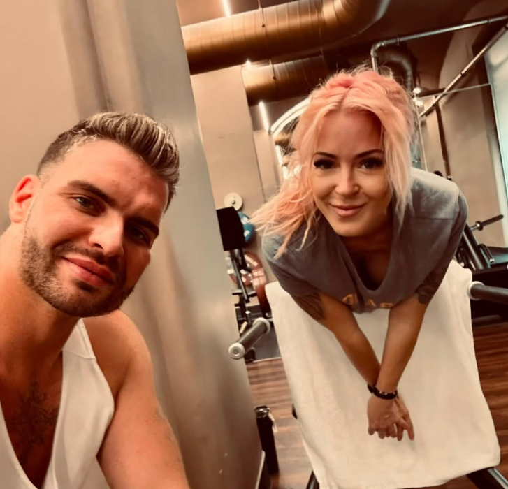 Ein Mann und eine Frau lächeln in einem Fitnessstudio in die Kamera.